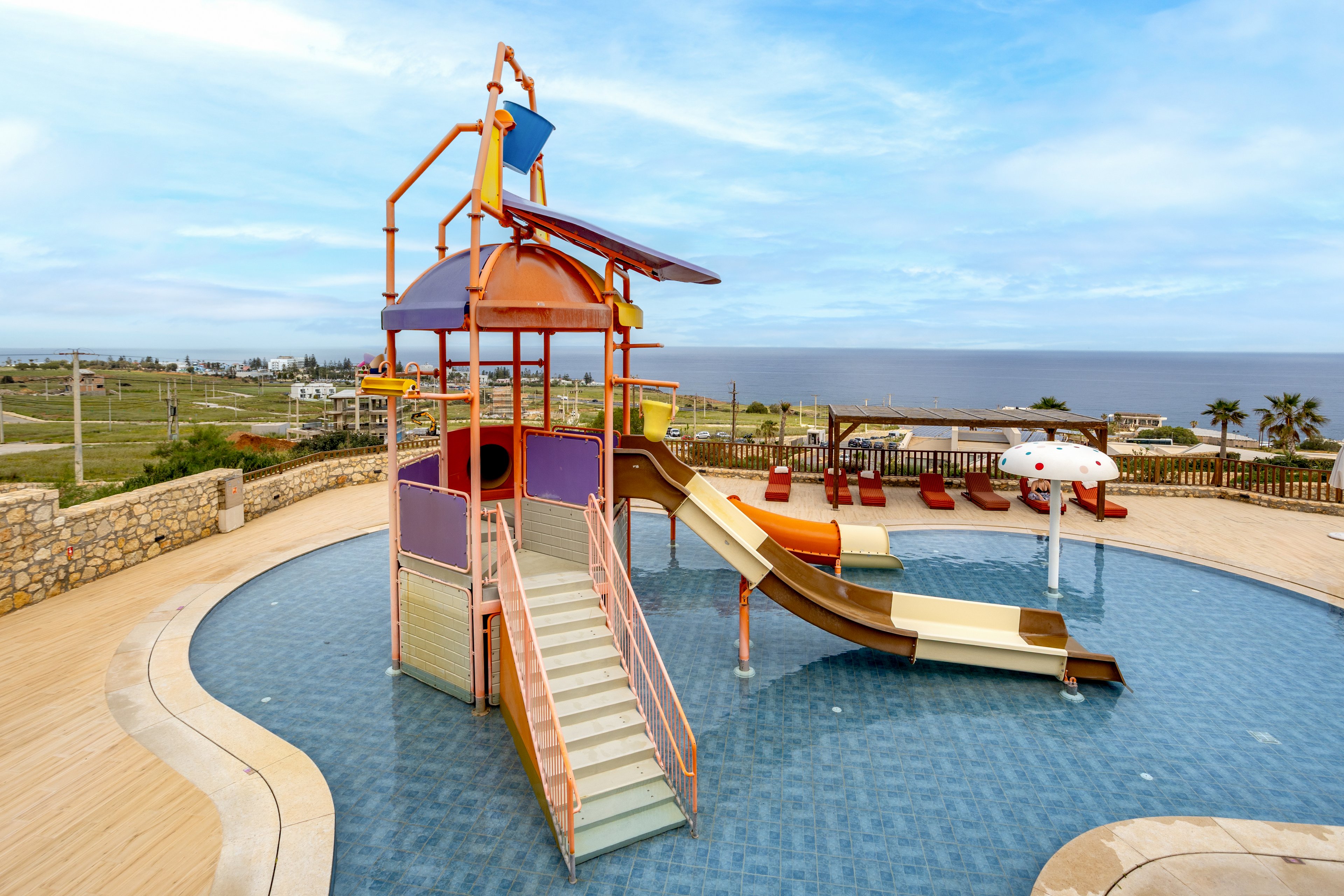 A brightly colored water playground with slides stands in the center of a shallow circular pool. The structure features multiple slides, platforms, and a small water mushroom feature. Lounge chairs and umbrellas line the poolside area, suggesting a family-friendly resort setting. In the background, open fields, palm trees, and a distant sea view create a relaxed vacation atmosphere.