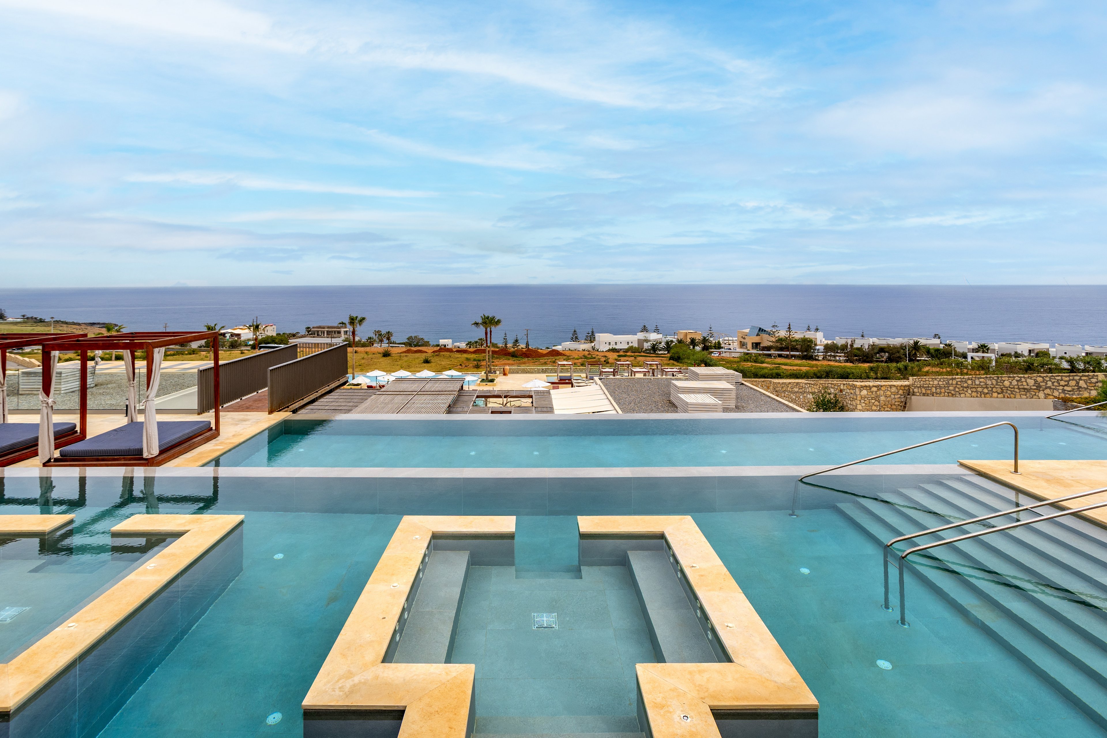 A modern multi-level infinity pool stretches toward a distant ocean horizon. Geometric hot tubs and clear blue water create a sleek, contemporary design. Beyond the pool area, a landscaped terrace and coastal buildings lead the eye to the calm sea. The bright sky and tranquil setting evoke a relaxing resort atmosphere.