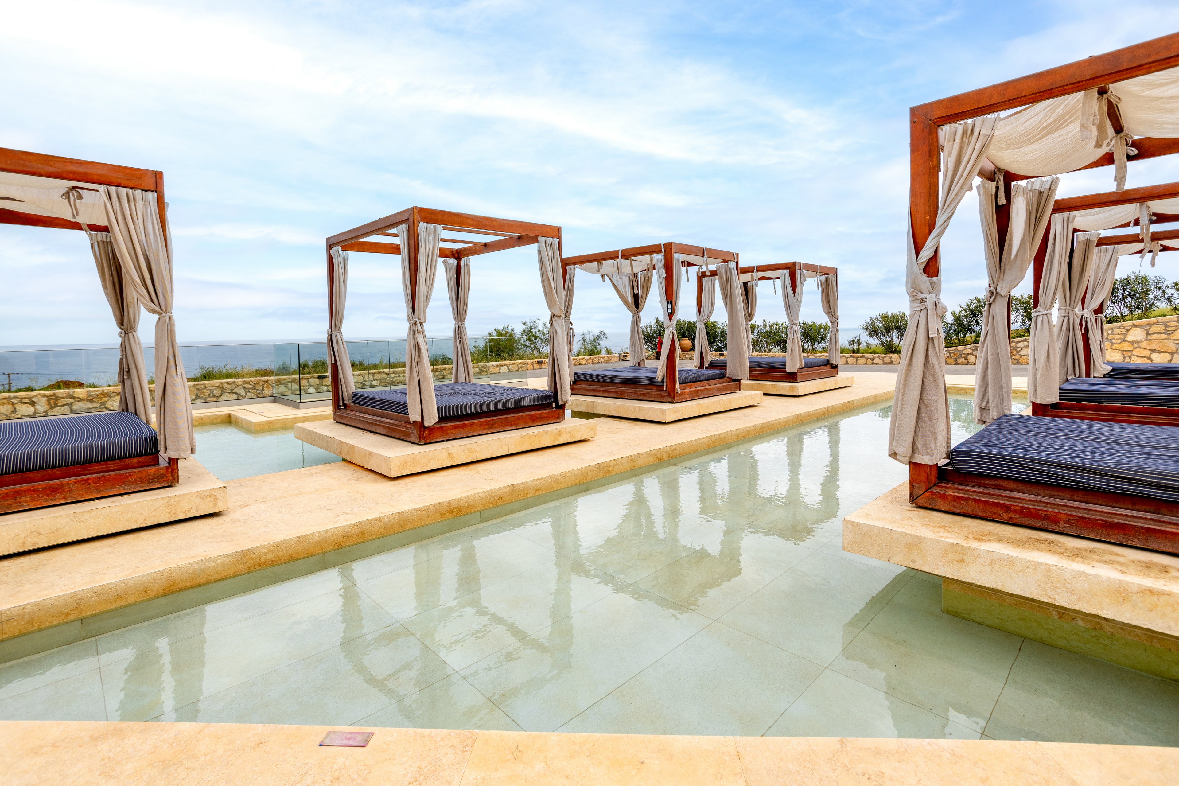 A row of modern wooden cabanas with draped white curtains stands beside a shallow reflective pool. Each cabana features dark cushioned daybeds arranged symmetrically along the water. The setting appears to be a high-end resort or spa with an ocean view in the background. Soft neutral tones of stone and fabric contrast with the blue sky and water, creating a calm, luxurious atmosphere.