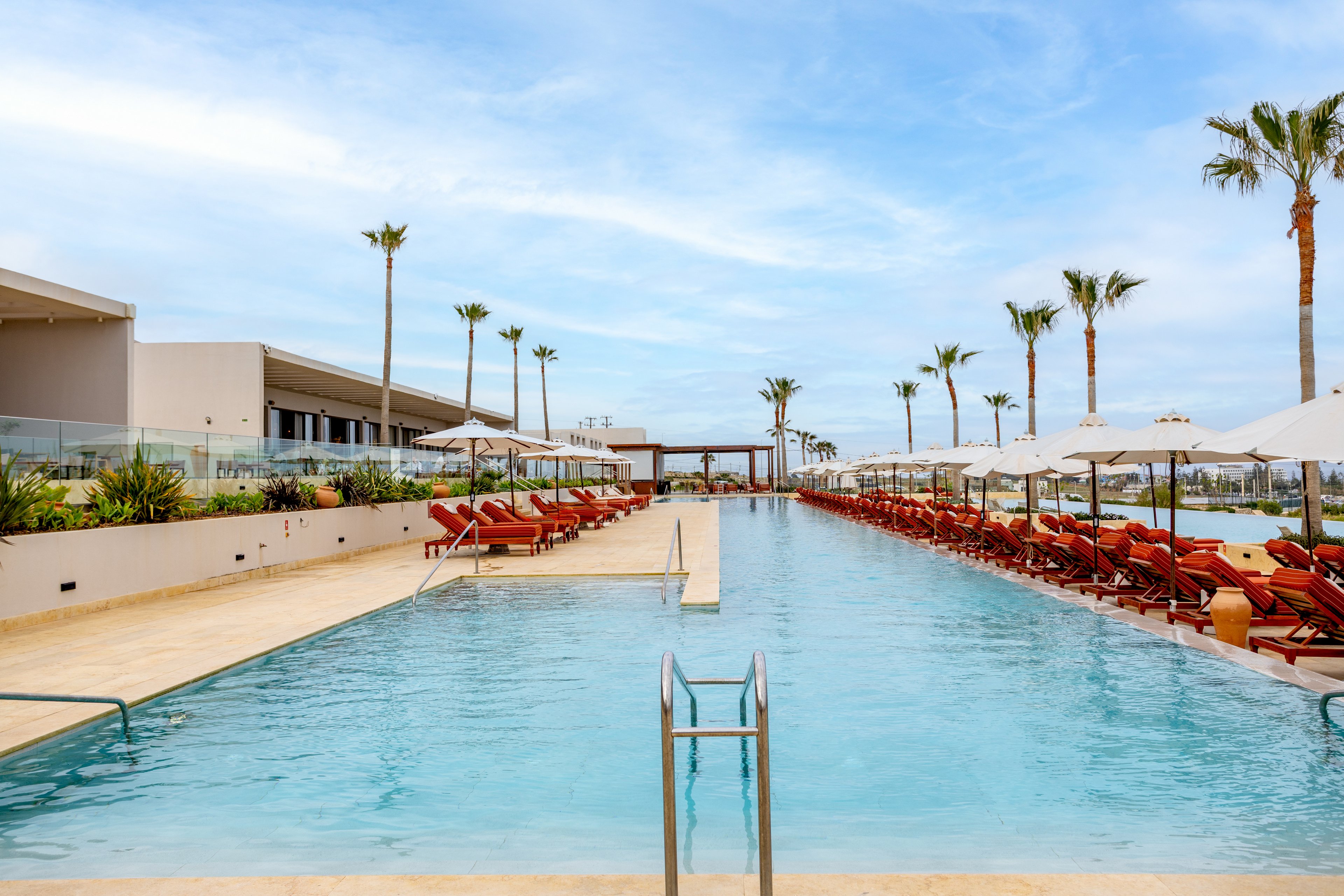 A long, modern infinity pool stretches between rows of red lounge chairs at a luxury resort. White umbrellas line both sides of the pool, providing shade along the sun deck. Tall palm trees rise above the contemporary buildings and glass railings in the background. The scene feels calm and inviting under a bright, partly cloudy sky.