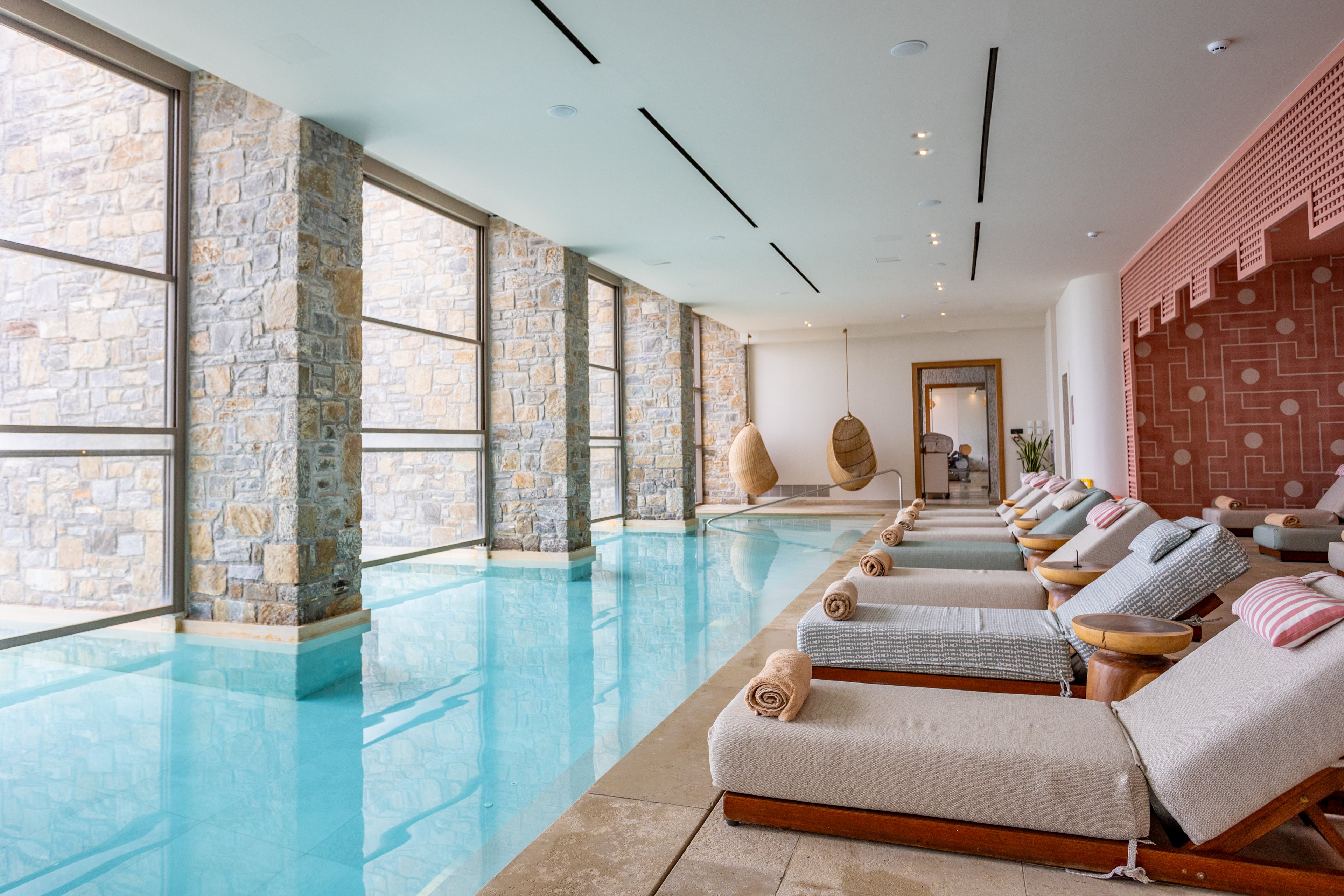 A serene indoor spa pool is lined with cushioned lounge chairs and neatly rolled towels. Large floor-to-ceiling windows with stone columns allow natural light to flood the space. Hanging wicker chairs are visible at the far end, adding a playful yet relaxing touch. The clean architectural lines and soft neutral tones create a calm, luxurious atmosphere ideal for wellness and relaxation.
