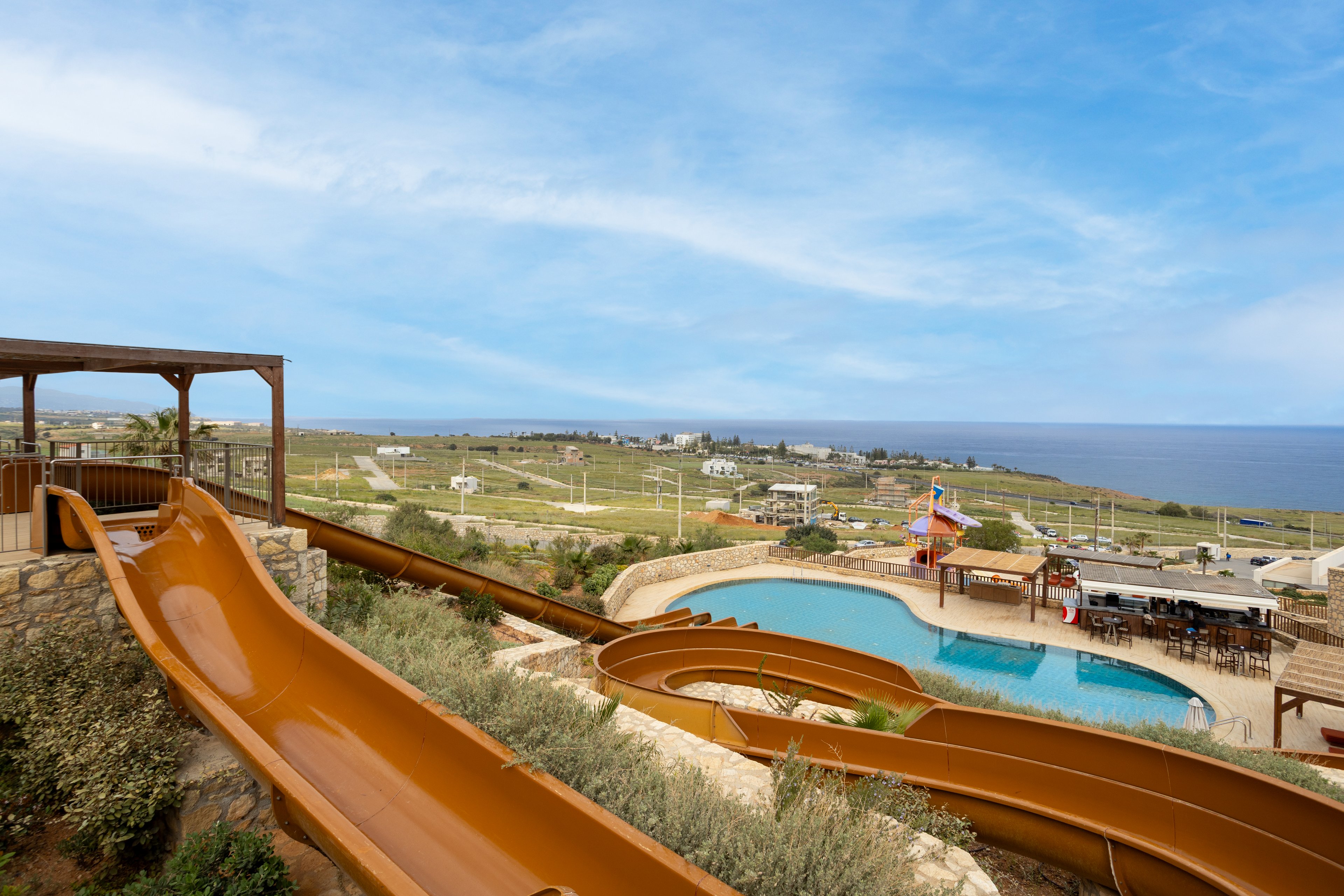 A pair of large brown waterslides curve down toward a resort swimming pool. The pool area includes sun loungers, umbrellas, and a small play structure with colorful slides. Beyond the resort, open fields and scattered buildings stretch toward the coastline. The calm blue sea and partly cloudy sky create a relaxed vacation atmosphere. The elevated viewpoint highlights both the waterpark features and the expansive coastal landscape.