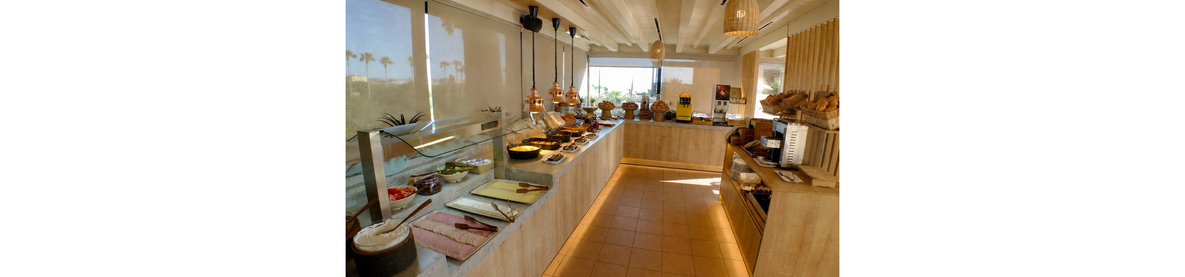 A spacious self-service buffet counter is set up with a variety of breakfast foods, including breads, pastries, cold cuts, and hot dishes in warming trays. The setting appears to be a modern hotel or resort dining area with light wood finishes and large windows. Natural daylight floods the room, and palm trees are visible outside, suggesting a warm, coastal location. The overall atmosphere is inviting and casual, ideal for guests enjoying a relaxed morning meal.