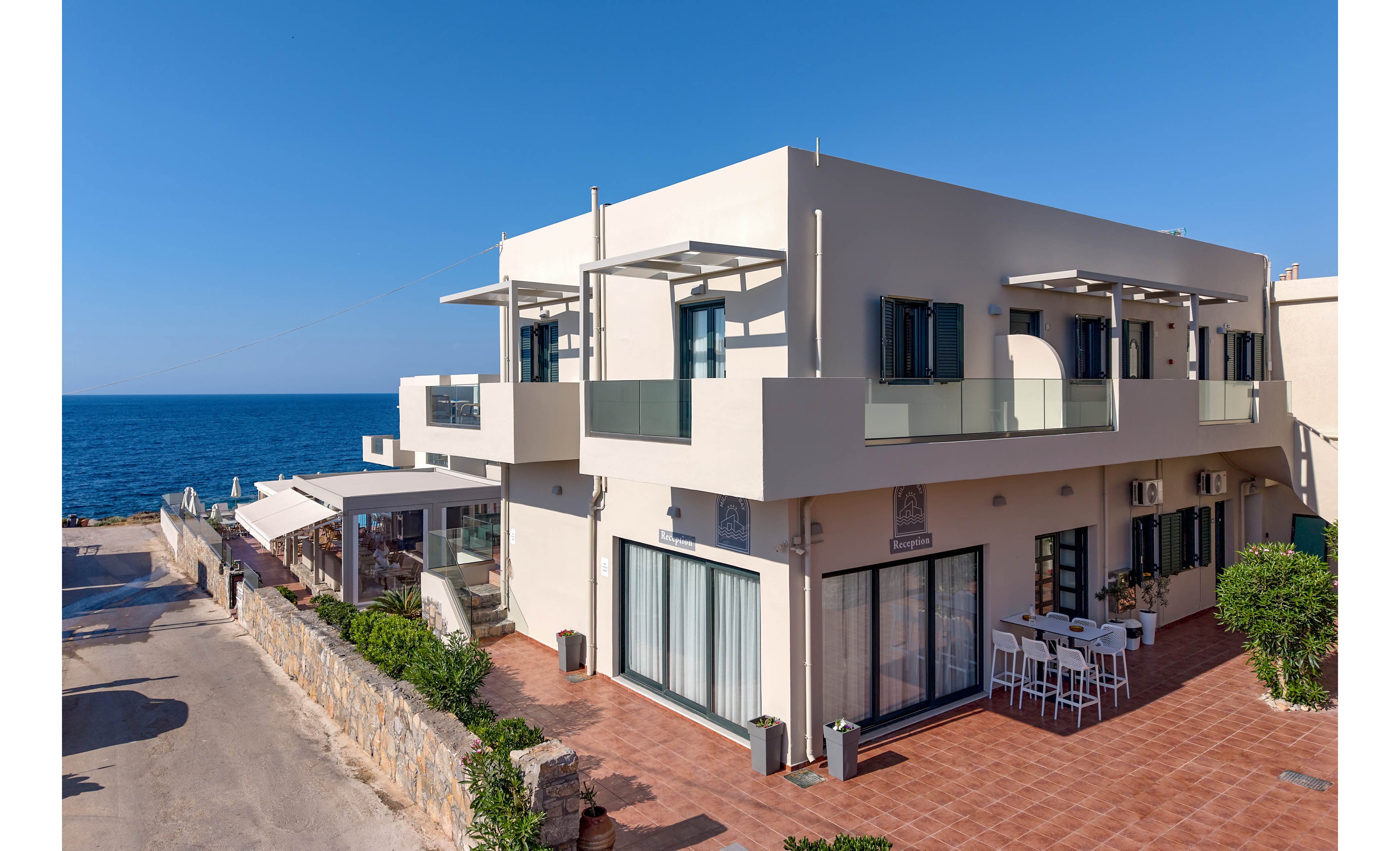 A modern two-story apartment building stands beside a coastal road overlooking the sea. The structure features light-colored walls, glass balconies, and large windows that open onto a spacious tiled terrace. Outdoor seating with white tables and chairs is arranged near the entrance, suggesting a café or communal area. The clear blue sky and calm water create a bright, relaxed Mediterranean atmosphere.