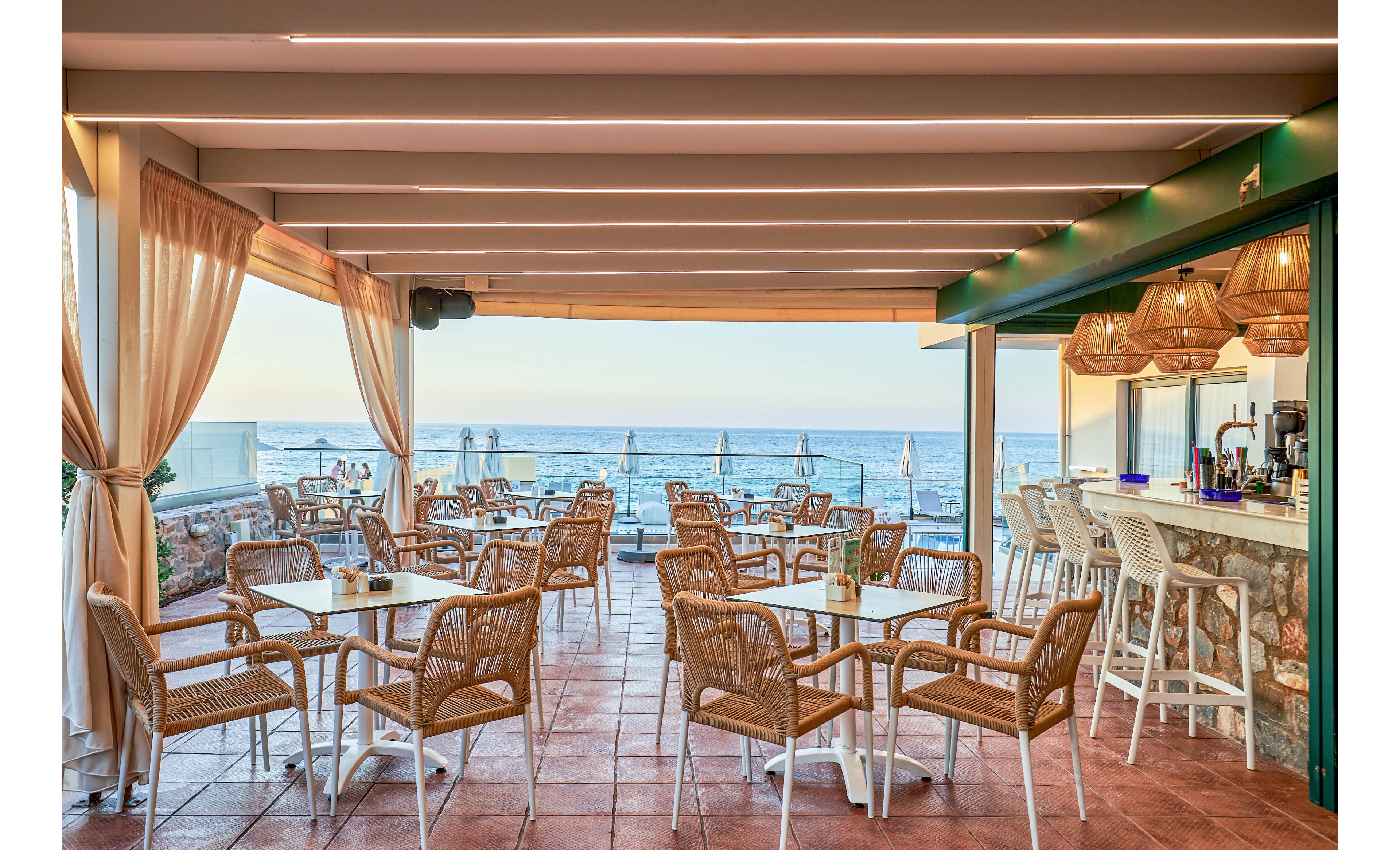 An open-air café with wicker chairs and small tables overlooks a beach and the ocean. The space is covered by a modern pergola with soft draped curtains and warm pendant lights above the bar area. Sun loungers and umbrellas are visible outside on the terrace near the shoreline. The scene feels relaxed and inviting, ideal for coastal dining or sunset drinks.