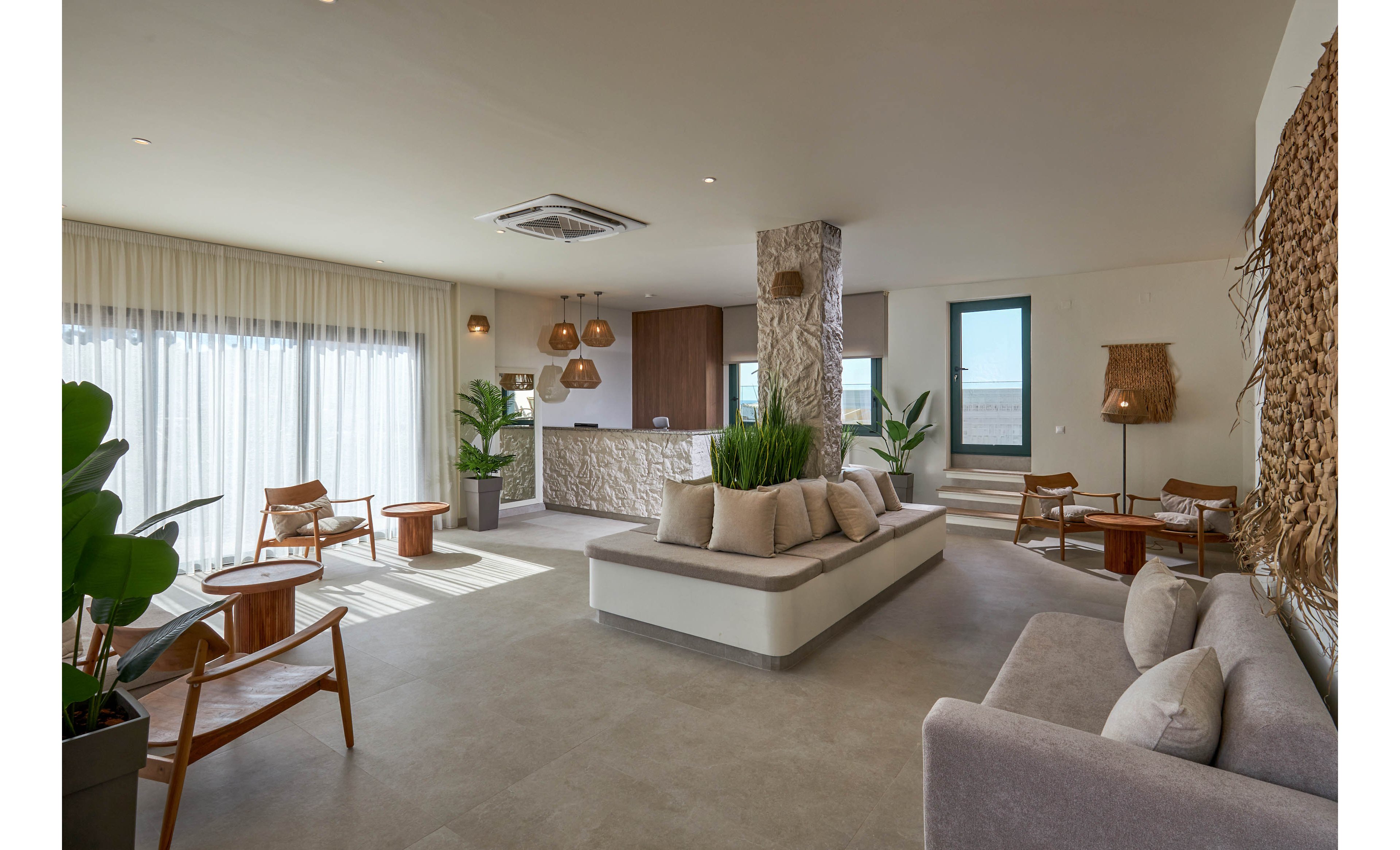 Spacious contemporary living room features minimalist furniture and neutral tones. Large floor-to-ceiling windows with sheer curtains fill the space with natural daylight. Wooden chairs, woven lamps, and indoor plants create a warm, organic atmosphere. A central cushioned bench and stone-clad column add texture and visual interest to the open-plan interior.