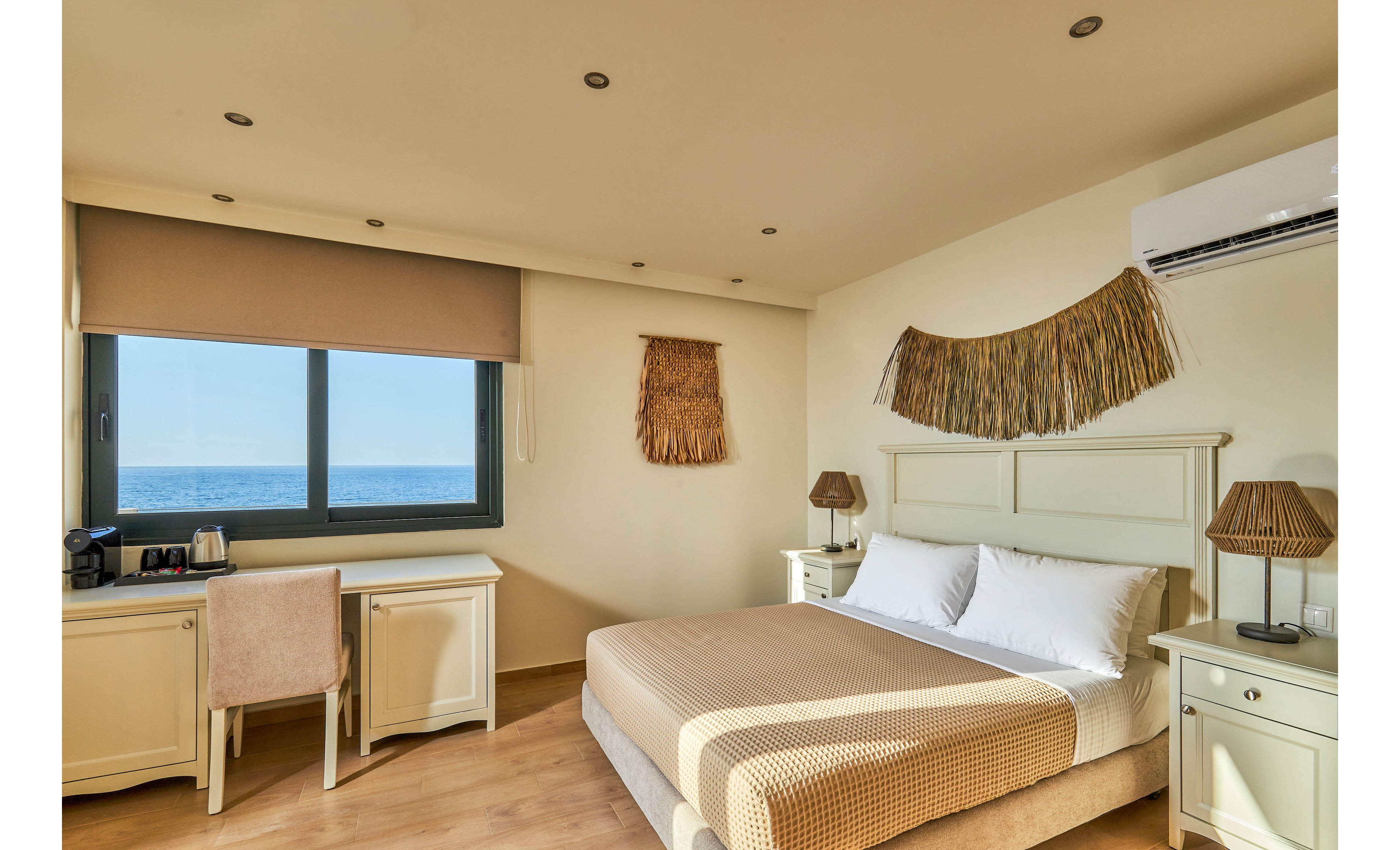 A sunlit hotel bedroom features a neatly made double bed with neutral bedding and matching bedside tables. A large window offers a clear view of the sea, filling the room with natural light. A desk and chair sit beside the window, equipped with a coffee machine and cups for guest convenience. Warm wooden flooring, woven wall decor, and soft beige tones create a calm, coastal atmosphere.