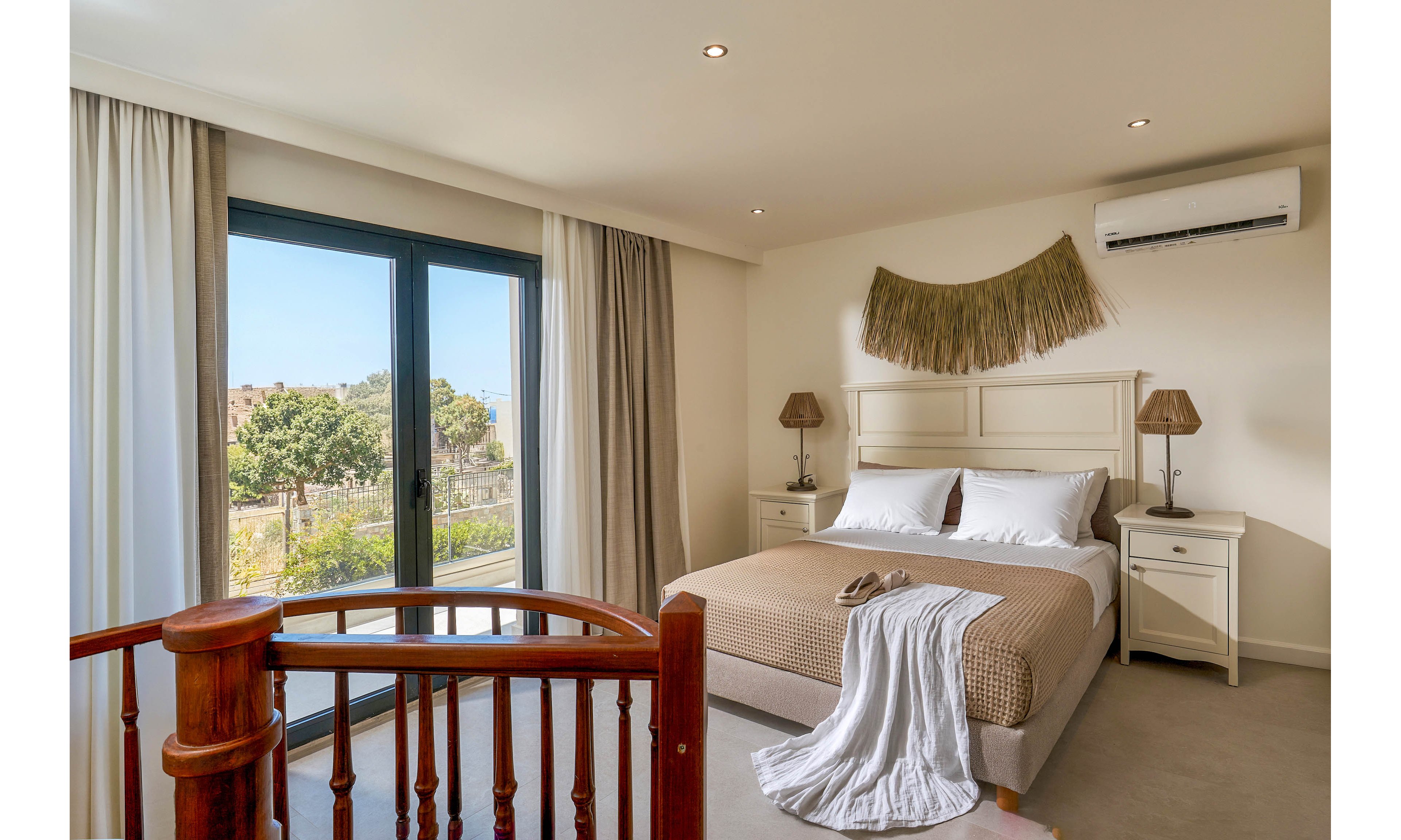 A contemporary bedroom features a double bed with neutral bedding and white pillows. Large glass doors open to a balcony with a scenic view of trees and buildings outside. The room includes bedside tables with lamps, wall-mounted air conditioning, and decorative wall art above the headboard. Soft natural light and light-colored curtains create a calm, inviting atmosphere.