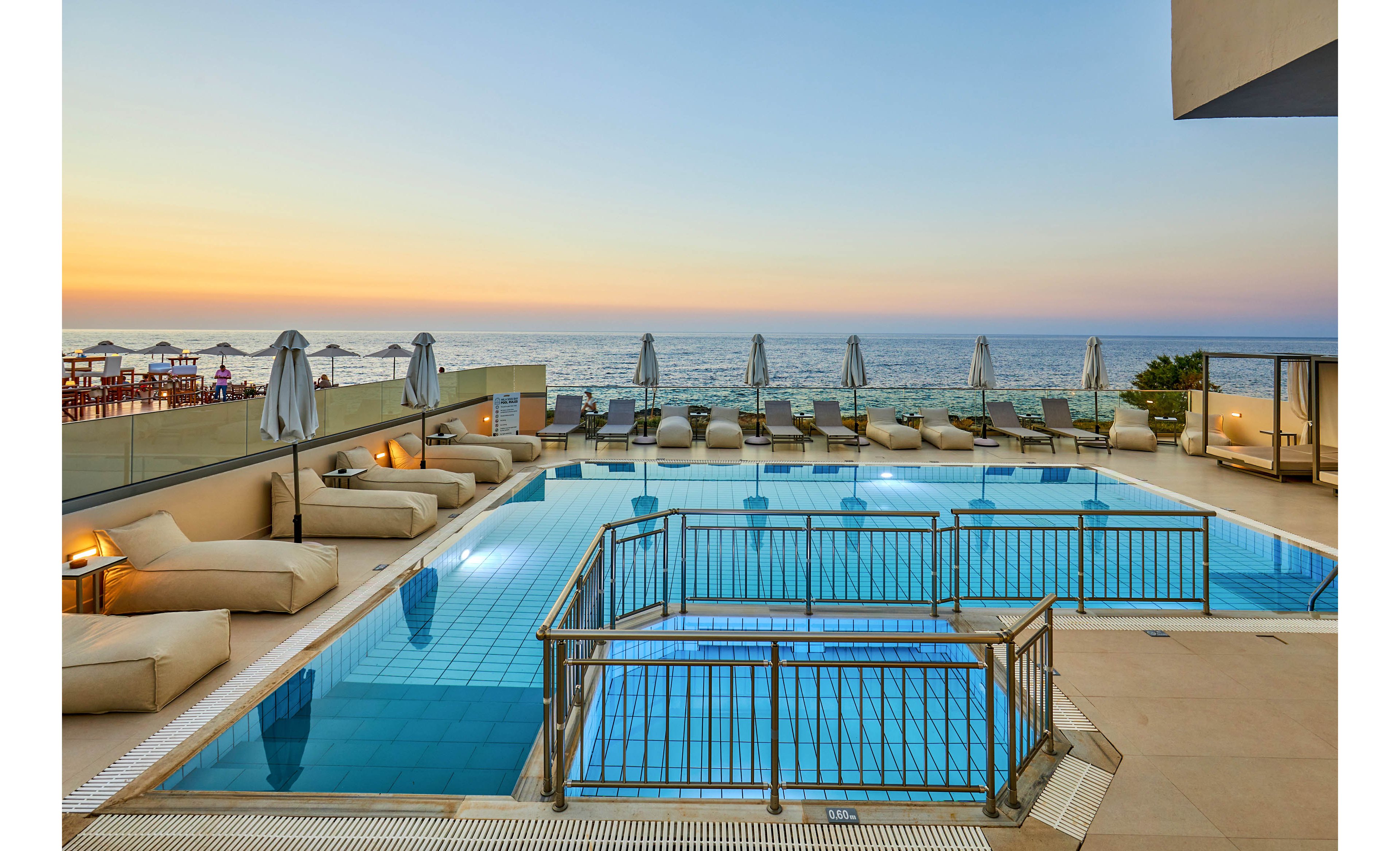 A modern outdoor infinity pool overlooks a calm sea at sunset, with soft pastel colors in the sky. Cushioned lounge beds and closed umbrellas line the poolside terrace, creating a relaxed resort atmosphere. Glass railings and clean architectural lines frame the water, reflecting the warm evening light. The scene suggests a tranquil vacation setting with ocean views and contemporary comfort.