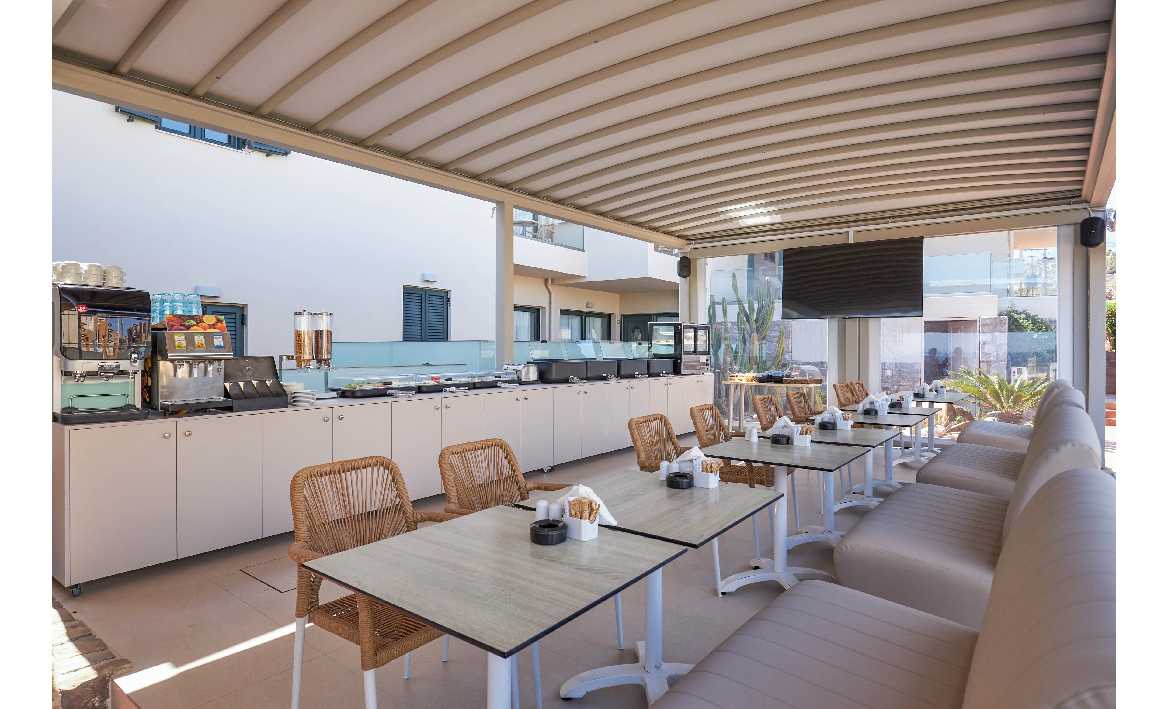 An open-air hotel café terrace features neatly arranged tables and cushioned bench seating under a modern pergola. Wooden chairs and light-colored furniture create a relaxed, contemporary atmosphere. In the background, a self-service counter with beverage dispensers and equipment lines the wall. Large windows and surrounding buildings suggest a resort or urban hotel setting with a clean, minimalist design.
