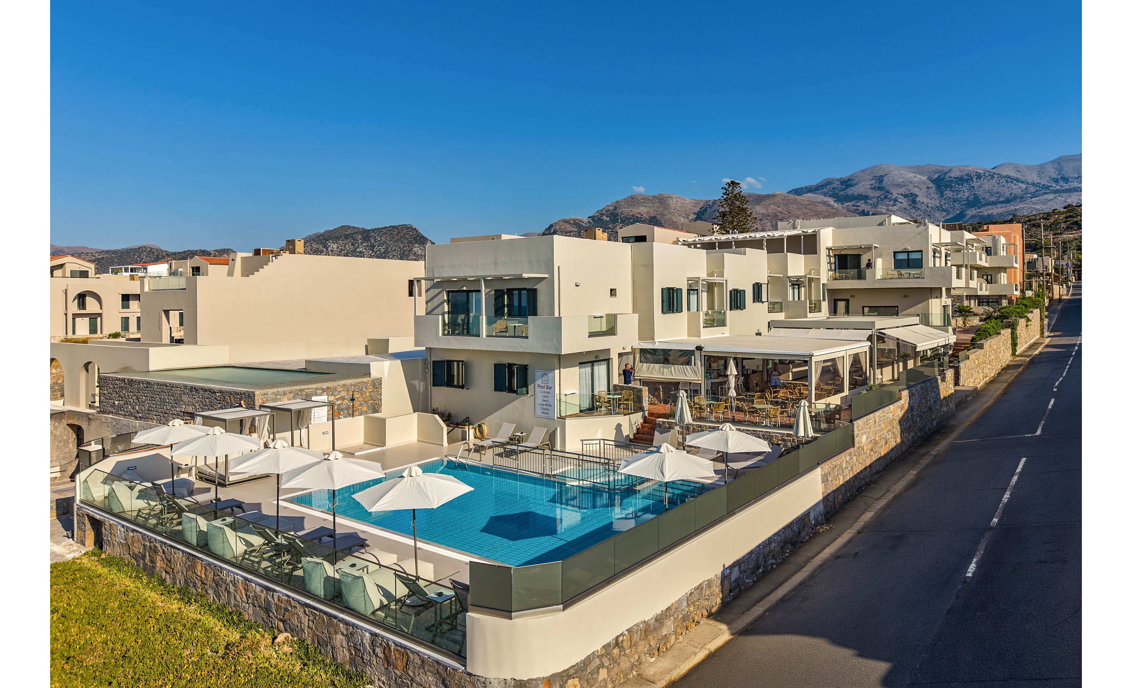 A contemporary resort complex features a central outdoor swimming pool surrounded by sun loungers and white umbrellas. The buildings have clean, modern architecture with balconies and terraces overlooking the pool area. The property sits beside a quiet paved road and is bordered by stone walls and glass railings. In the background, rugged mountains rise under a clear blue sky, adding a scenic natural backdrop to the vacation setting.