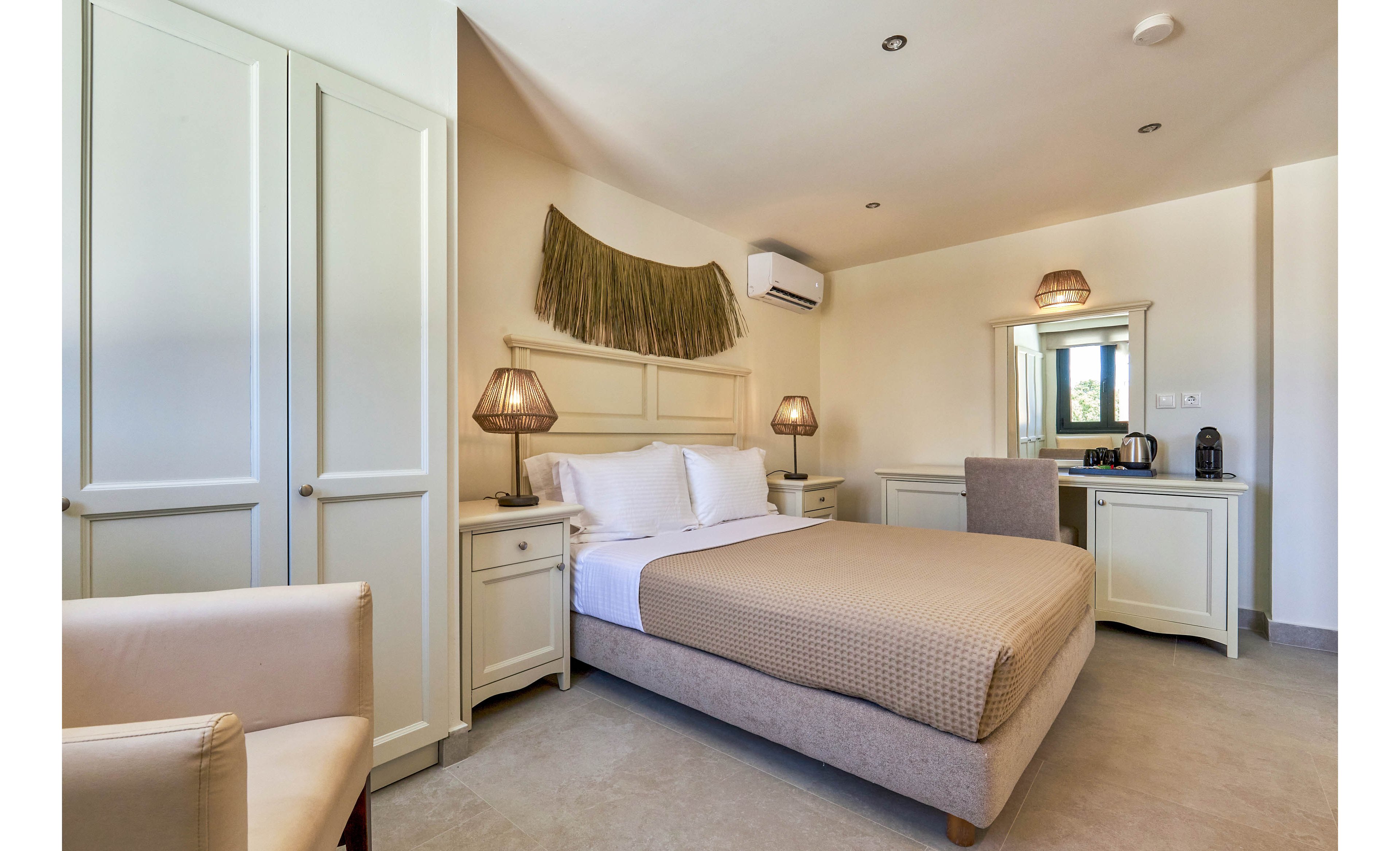 A neatly made double bed sits at the center of a bright, modern hotel bedroom with neutral tones. Matching bedside tables with woven lamps flank the bed, and a decorative wall hanging adds texture above the headboard. A wardrobe and upholstered chair are visible on one side, while a desk with mirror, electric kettle, and coffee machine occupies the opposite wall. An air conditioning unit is mounted above, and natural light enters through a window reflected in the mirror.