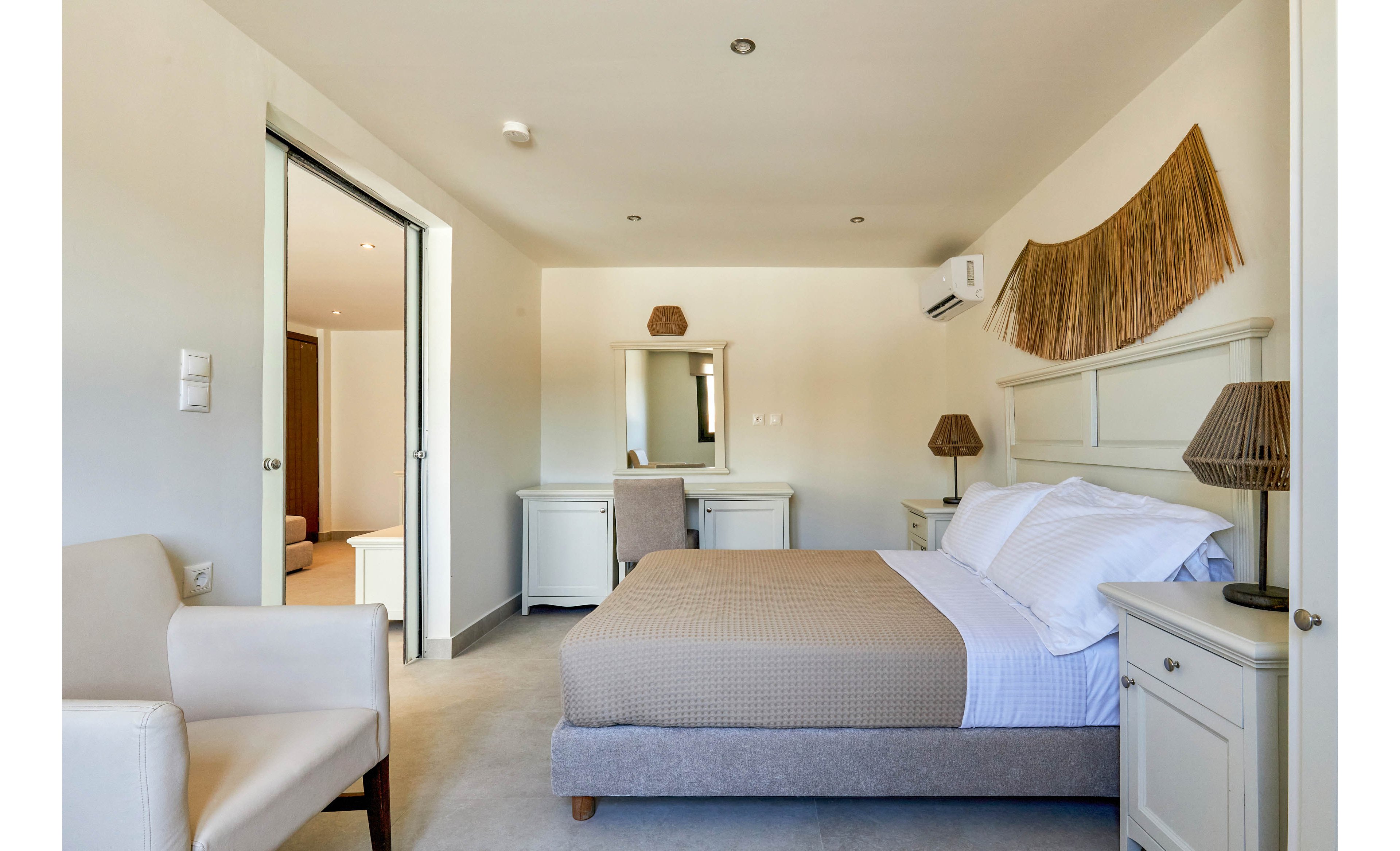A contemporary bedroom features a double bed with white linens and a beige textured blanket. Matching bedside tables and lamps flank the bed, while a vanity with mirror and chair sits against the far wall. An armchair is positioned near the doorway, and an open sliding door reveals an adjacent sitting area. The room is decorated in soft neutral tones with minimalist furnishings and clean lines.