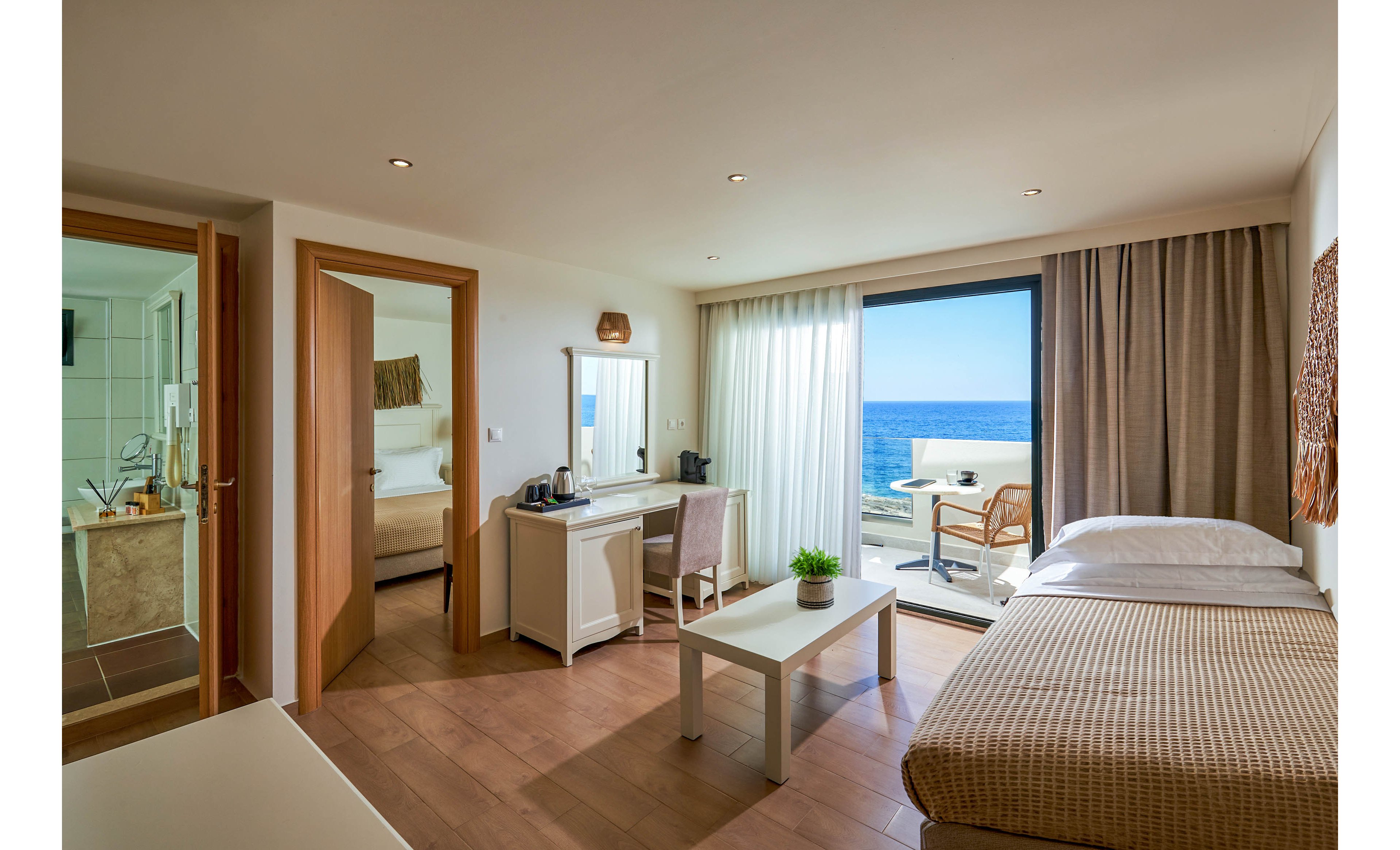A modern hotel room features a single bed, a small table with a potted plant, and a vanity desk with a mirror and chair. Large sliding glass doors open onto a balcony with chairs, revealing a clear view of the ocean and blue sky. Warm wood tones, neutral textiles, and soft lighting create a calm, inviting atmosphere. An adjacent bathroom and secondary sleeping area are visible through open doors, adding depth to the interior space.