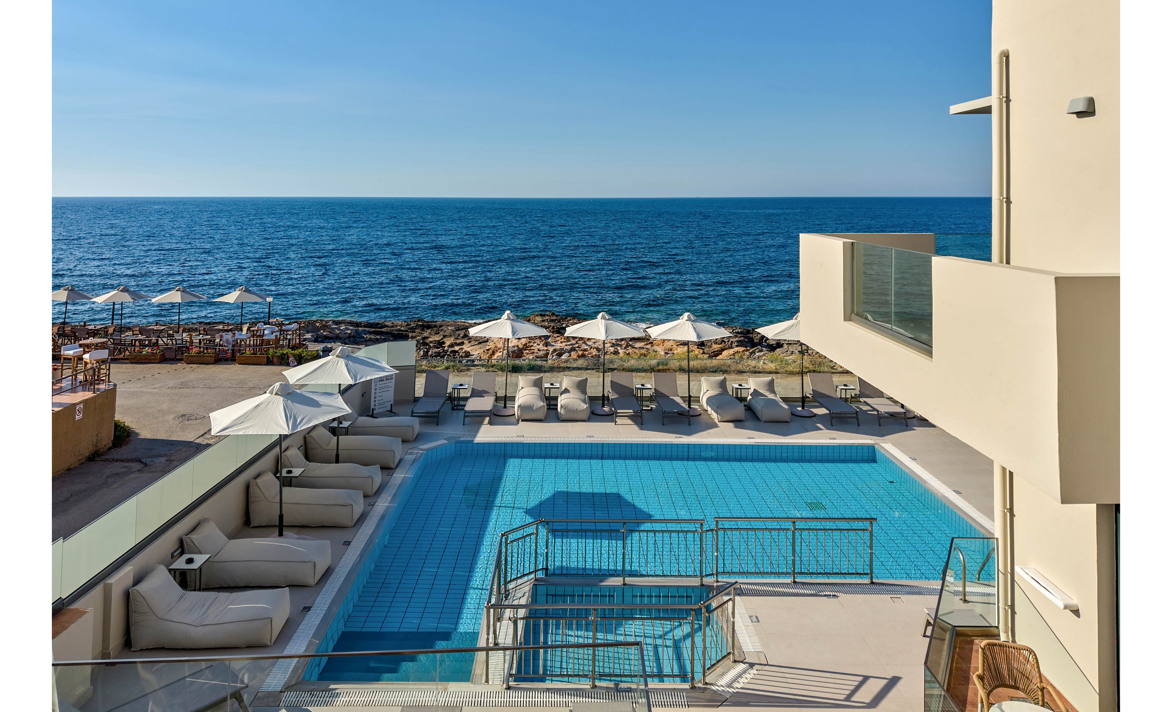 A modern hotel pool overlooks a calm blue ocean under a clear sky. White sun loungers and umbrellas line the poolside terrace, creating a relaxed resort atmosphere. Beyond the pool area, additional seating and umbrellas sit near a rocky shoreline. Clean architectural lines and neutral tones contrast with the vivid blue water, emphasizing a contemporary coastal design.
