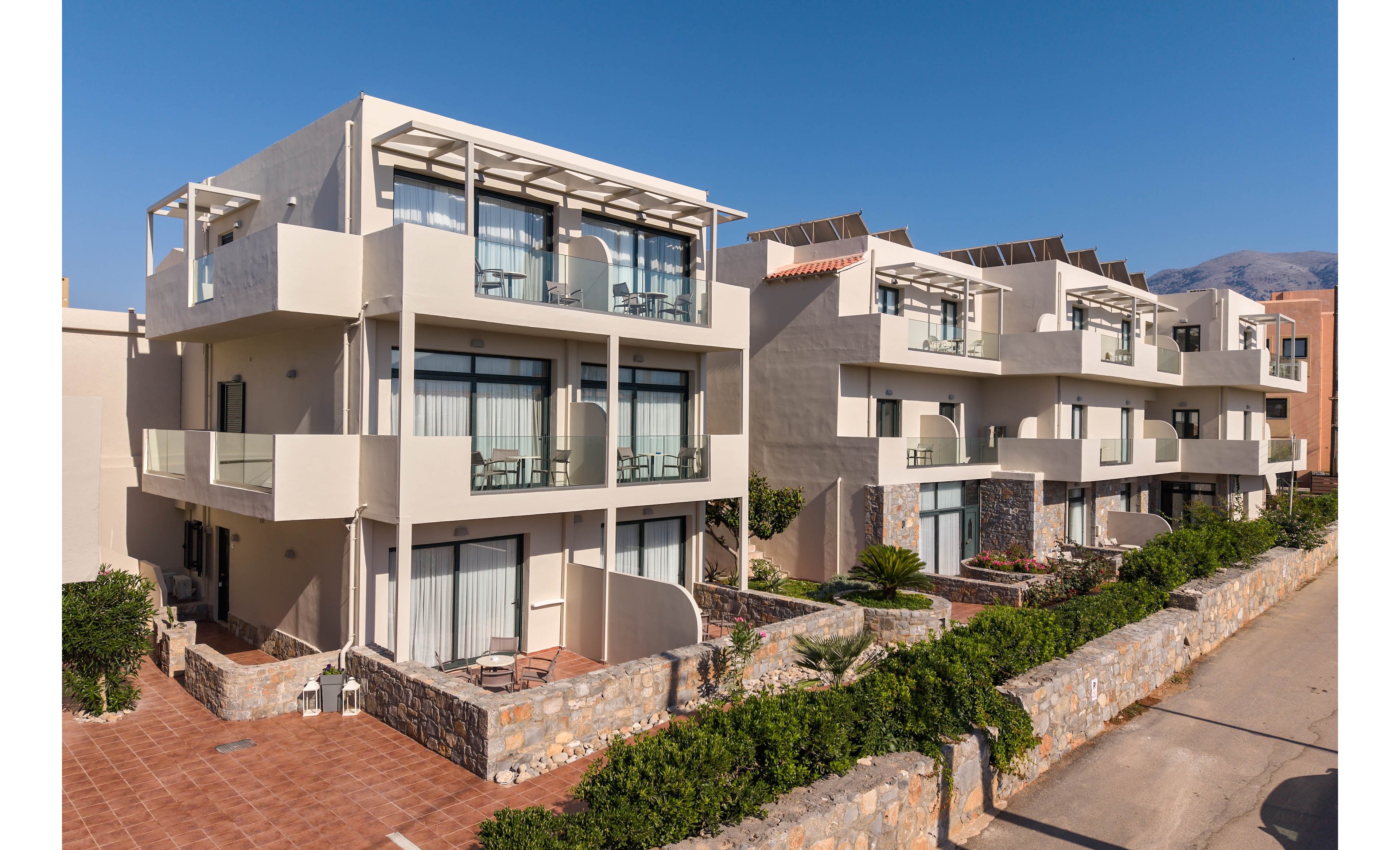 The image shows a row of modern multi-story apartment buildings with large glass balconies and terraces. The structures feature light-colored walls, stone accents, and neatly maintained ground-level patios. A narrow street runs alongside the buildings, bordered by low stone walls and greenery. In the background, distant hills or mountains are visible under a clear blue sky, suggesting a coastal or resort setting.