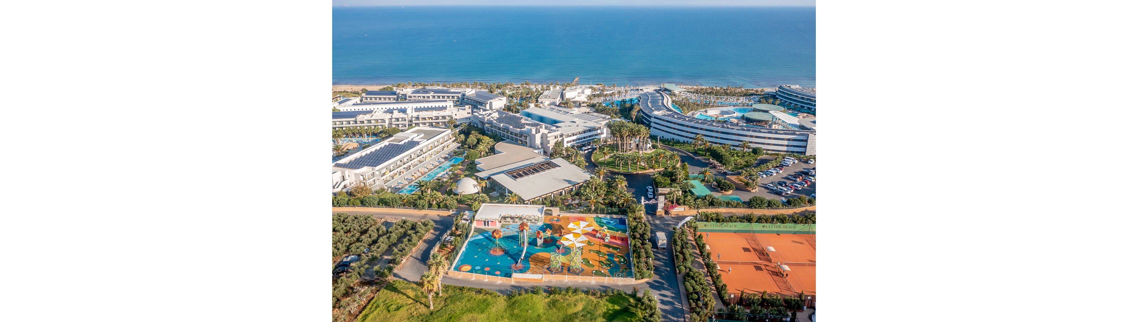 An expansive beachfront resort featuring multiple buildings, pools, and a vibrant water park with colorful designs. The setting includes lush greenery, tennis courts, and direct access to the ocean. The aerial perspective highlights the resort's layout and proximity to the coastline.