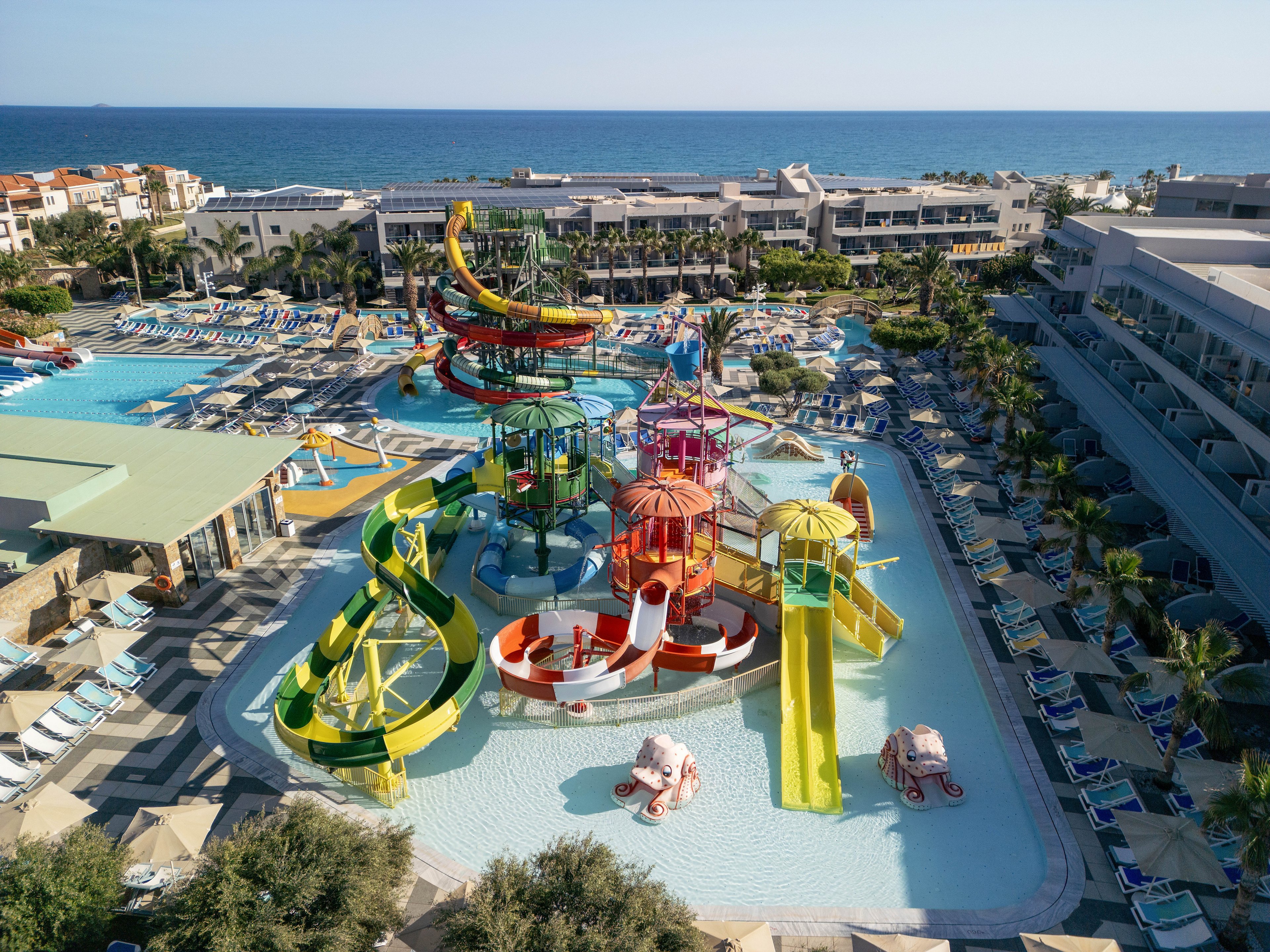 A large resort water park features multiple colorful slides and play structures surrounded by pools. The complex is located near the sea, with the ocean visible in the background beyond the hotel buildings. Numerous lounge chairs and umbrellas line the pool decks, suggesting a busy vacation destination. The bright colors of the slides contrast with the blue water and clear sky, creating a lively, family-friendly atmosphere.