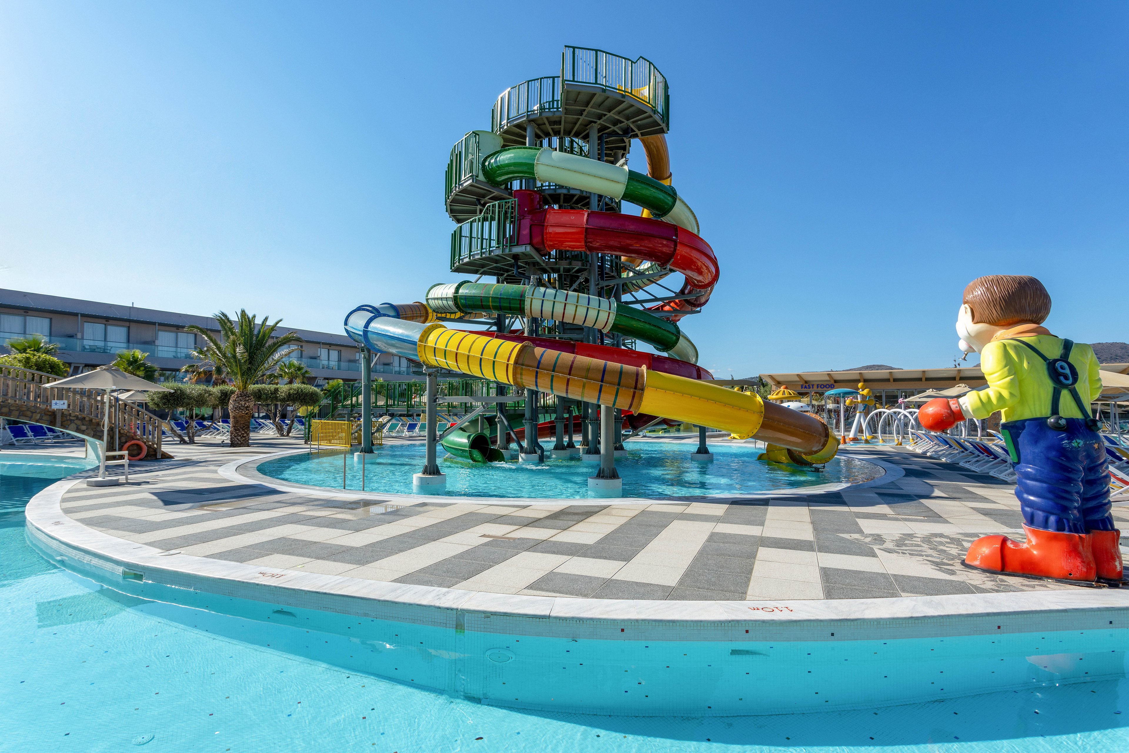 A large multi-level spiral waterslide tower rises above a shallow resort swimming pool. Several brightly colored enclosed and open slides twist down into the clear blue water. A large cartoon-style figure stands at the pool edge, adding a playful theme-park feel. The scene is set outdoors under a clear blue sky with lounge areas and palm trees in the background, suggesting a sunny vacation atmosphere.