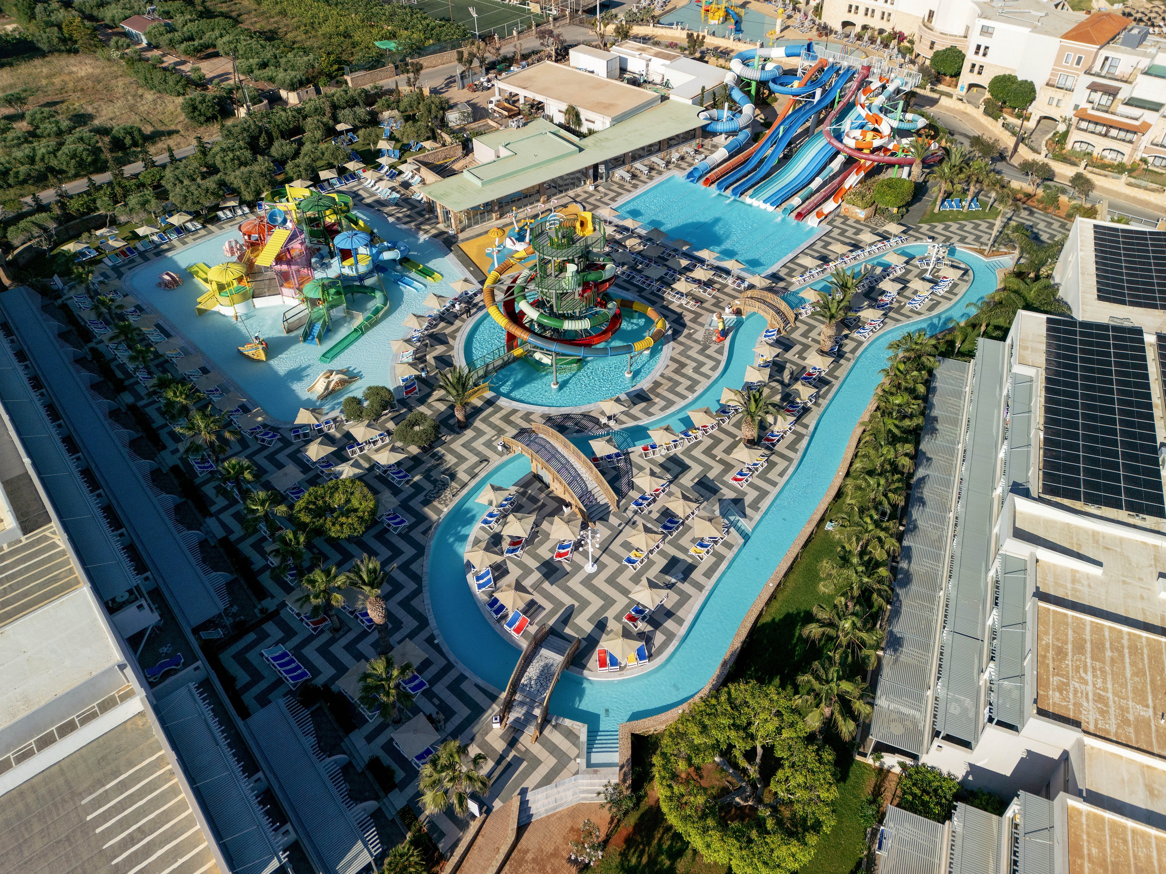 A large resort-style water park is captured from above, showing multiple pools, winding lazy rivers, and colorful water slides. Sun loungers and umbrellas line the pool decks, creating a structured pattern around the water features. A central play area with fountains and small slides is visible, surrounded by palm trees and landscaped paths. Buildings and green spaces frame the complex, emphasizing the scale of the recreational facility.