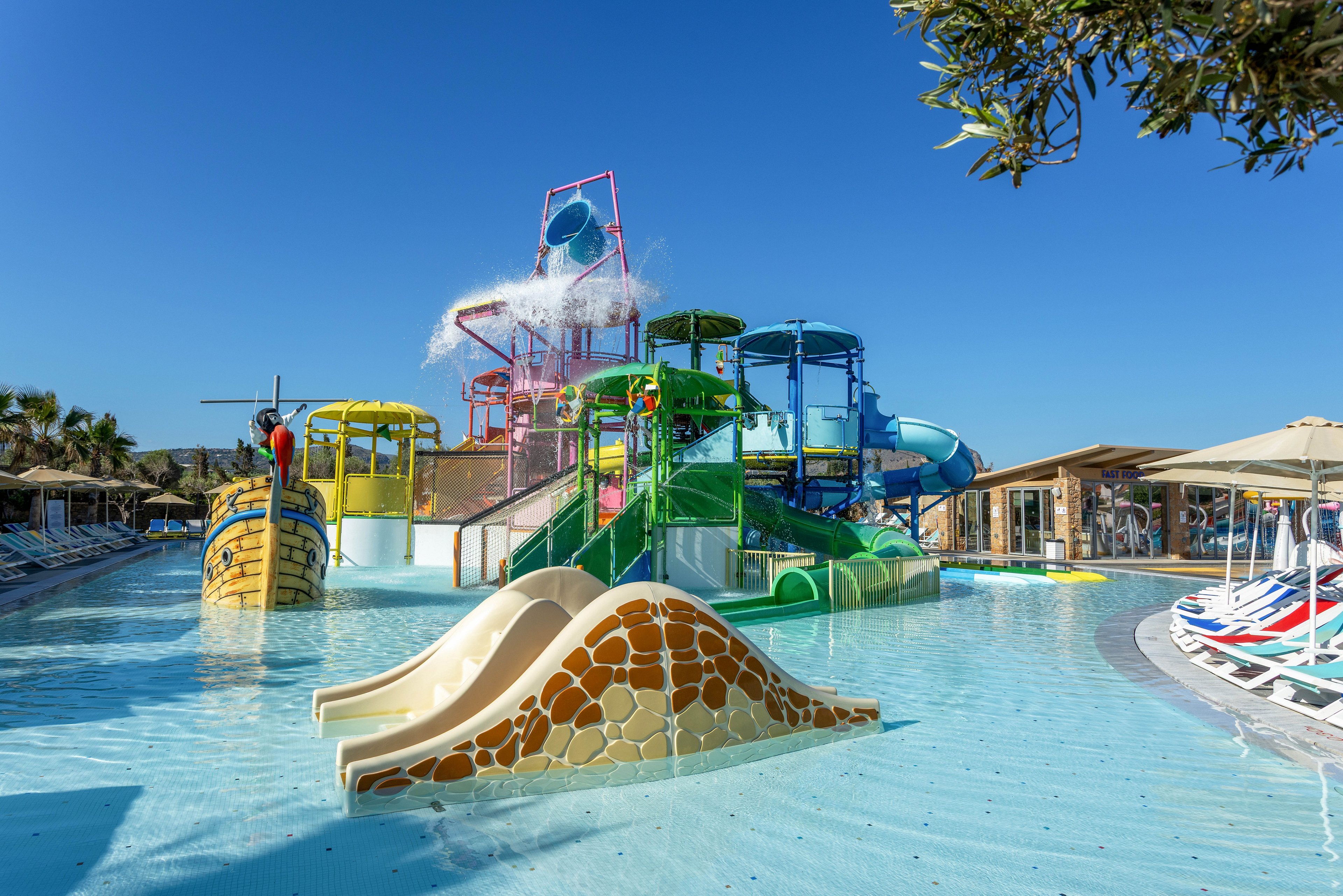 A vibrant outdoor water park features multiple colorful slides and play structures in a shallow pool. A large tipping bucket pours water over the central climbing frame, creating a splash under the clear blue sky. The scene includes themed elements like a ship-shaped structure and gently curved slides in the foreground. Sun loungers and umbrellas line the edges of the pool, suggesting a family-friendly resort setting.