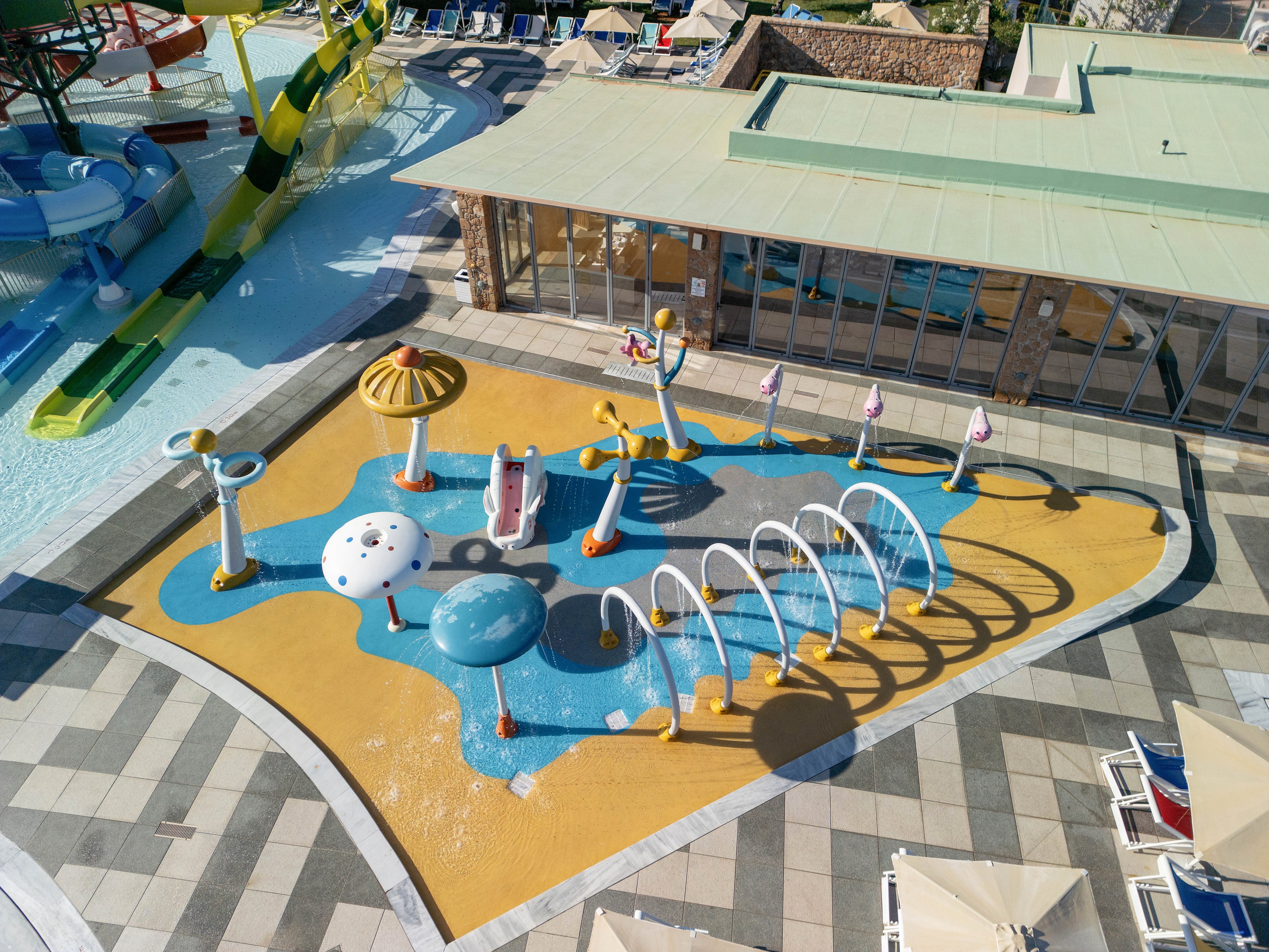 A brightly colored children's splash pad is seen from above in an outdoor water park. The play area features fountains, small slides, arches spraying water, and mushroom-shaped sprayers on a yellow and blue rubber surface. Surrounding the splash zone are tiled walkways, lounge chairs, and a nearby building with large windows. Adjacent pools and water slides indicate a larger aquatic recreation complex in a sunny resort setting.