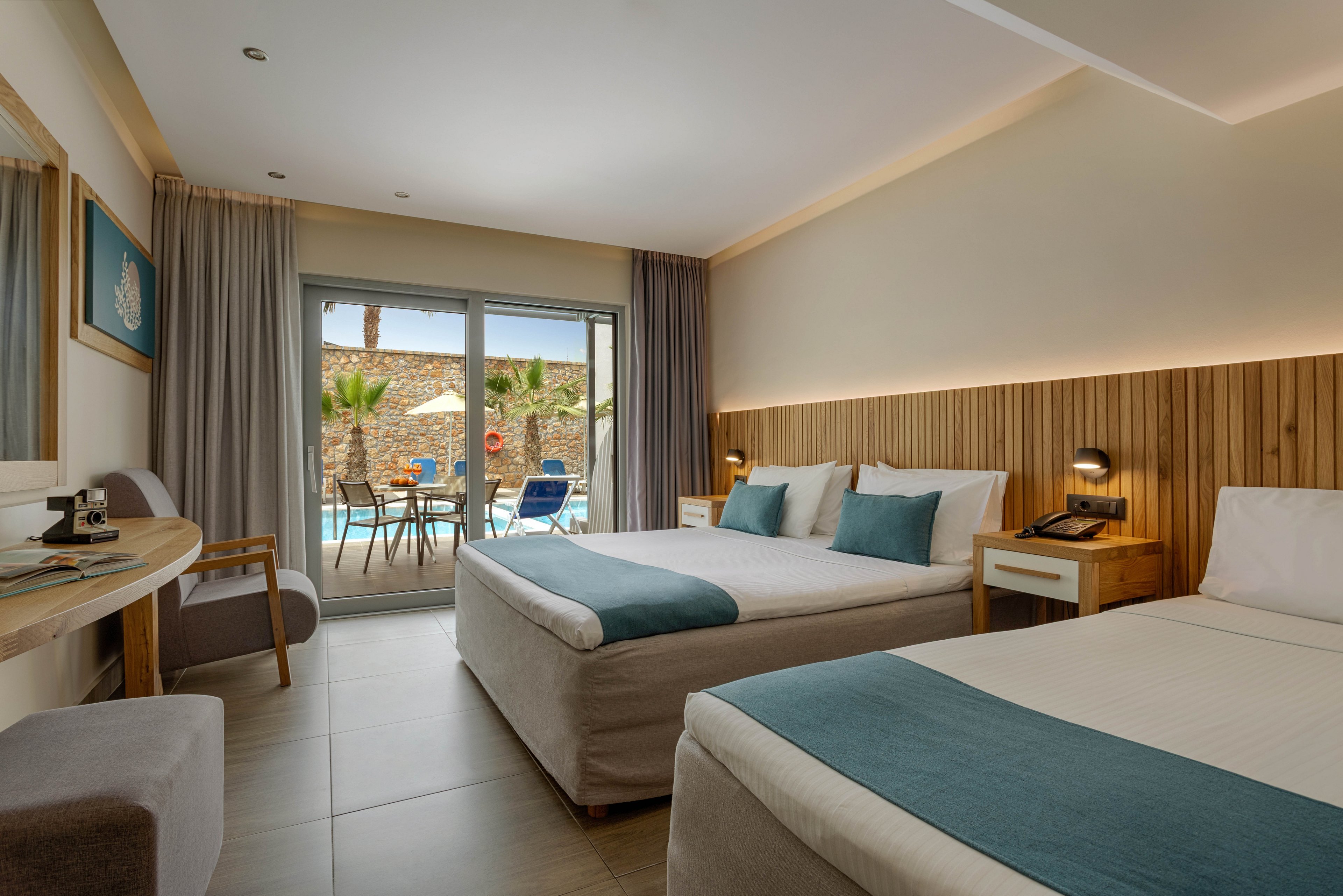 A contemporary hotel room features two neatly made double beds with teal runners and cushions. The space includes a wooden desk, bedside tables, and soft neutral furnishings that create a calm, inviting atmosphere. Large sliding glass doors open to a private terrace with chairs and a table overlooking a swimming pool and palm trees. Natural light fills the room, highlighting the minimalist decor and clean lines.