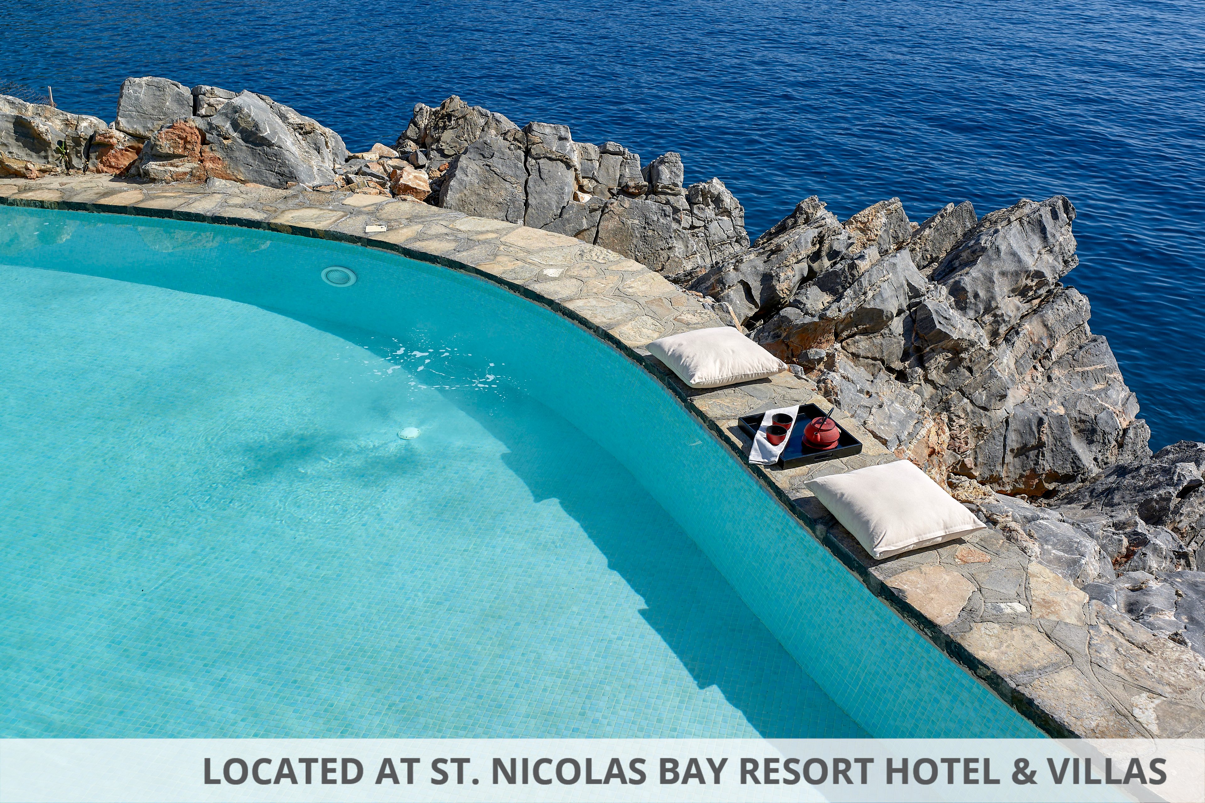 An infinity-edge swimming pool curves along a rocky shoreline, blending seamlessly with the blue ocean beyond. Two white cushions and a tray with drinks rest on the stone poolside, suggesting a relaxing seaside retreat. The clear turquoise water contrasts with the rugged gray rocks and deep blue sea. Bright daylight and calm conditions create a tranquil, luxurious vacation atmosphere.