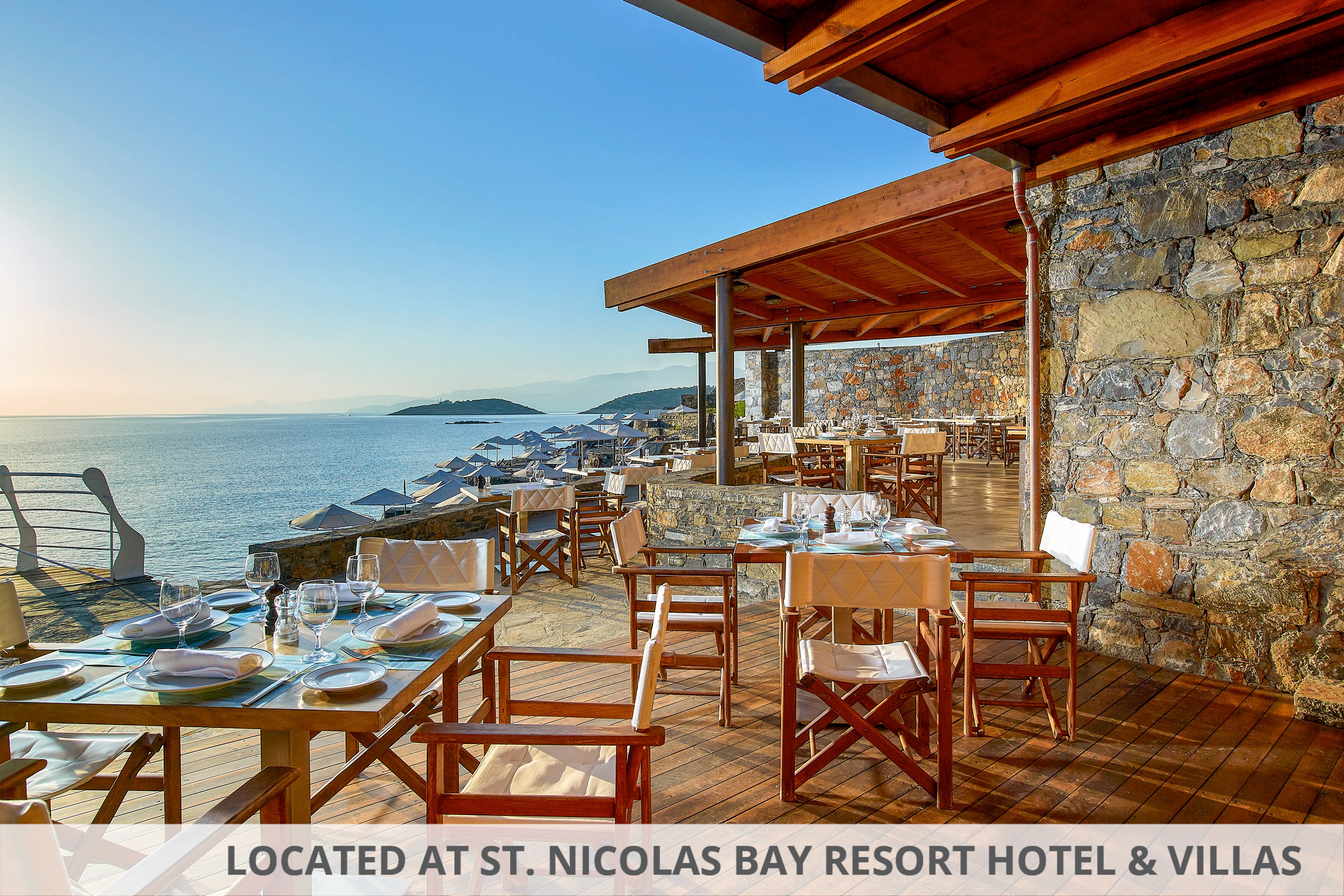 An open-air restaurant terrace with wooden tables and chairs overlooks a calm, blue ocean. Sunlight illuminates neatly set tables with glassware and plates, ready for guests. Stone walls and wooden decking create a rustic yet elegant coastal atmosphere. In the distance, lounge chairs line the waterfront and a small island is visible on the horizon under a clear sky.