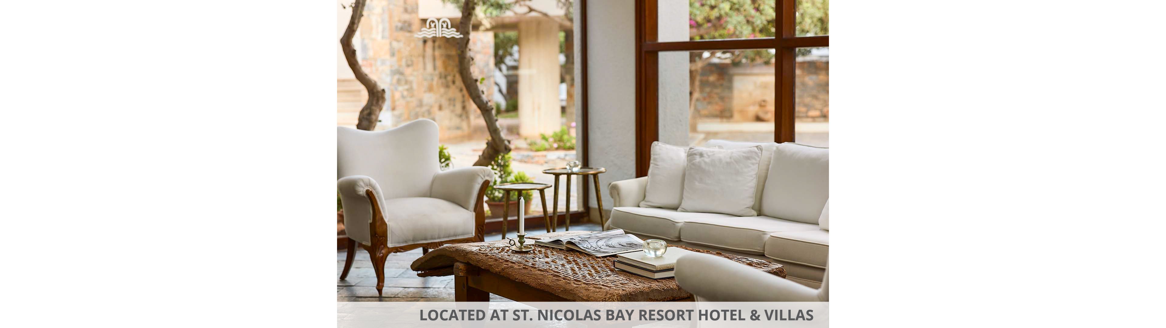 A cozy lounge area features a white sofa and matching armchairs arranged around a rustic wooden coffee table. Large windows open to a stone patio and trees, filling the room with natural light. A stack of books and a magazine rest on the textured table, adding a relaxed, lived-in feel. Neutral tones and soft upholstery create a calm, inviting atmosphere.