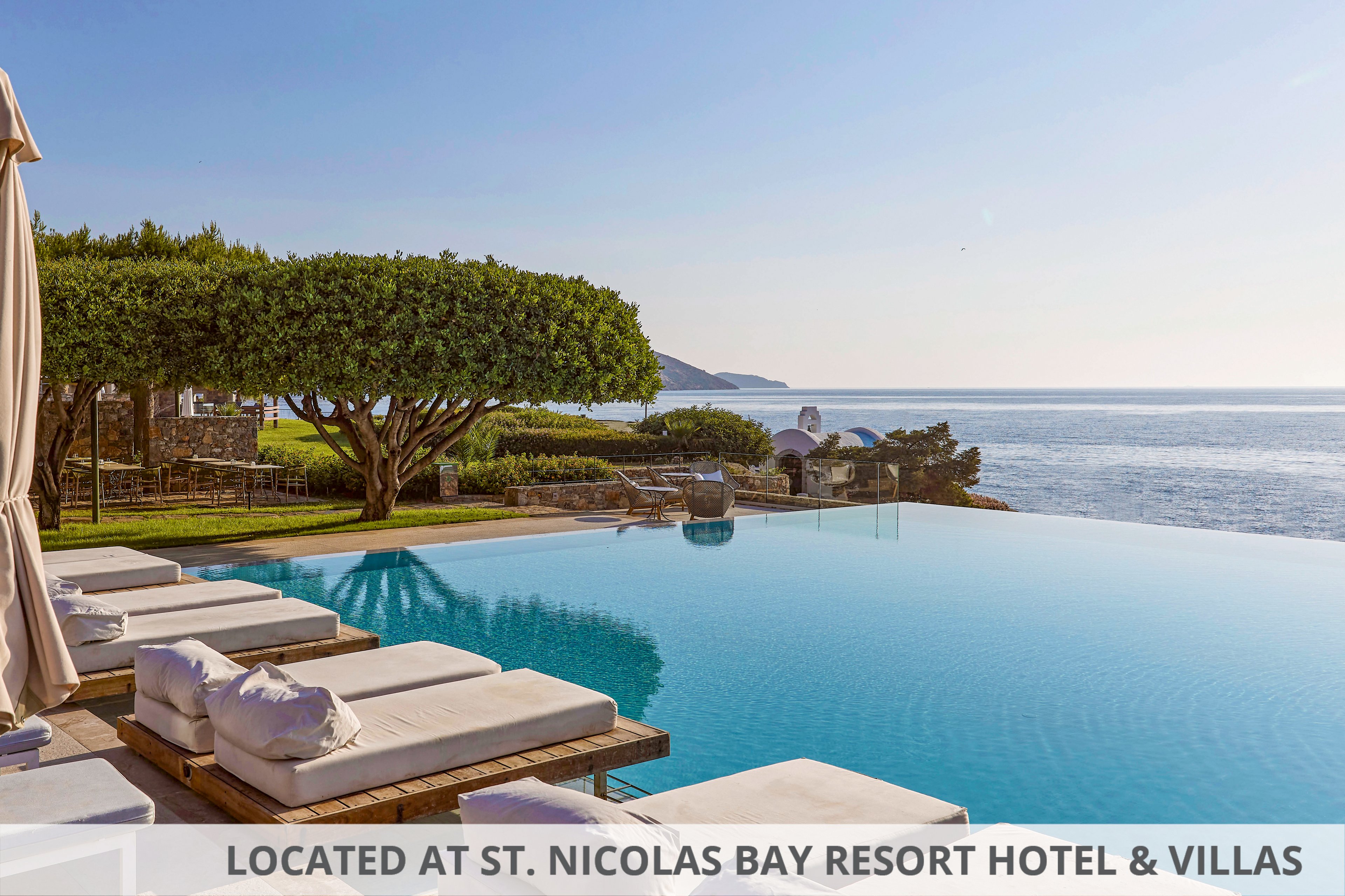 A serene infinity pool stretches toward a calm, expansive sea under a clear sky. White cushioned loungers line the poolside, suggesting a luxurious resort setting. Manicured trees and greenery frame the scene, while distant hills and coastline fade into the horizon. The overall atmosphere is peaceful and inviting, ideal for relaxation and coastal vacations.