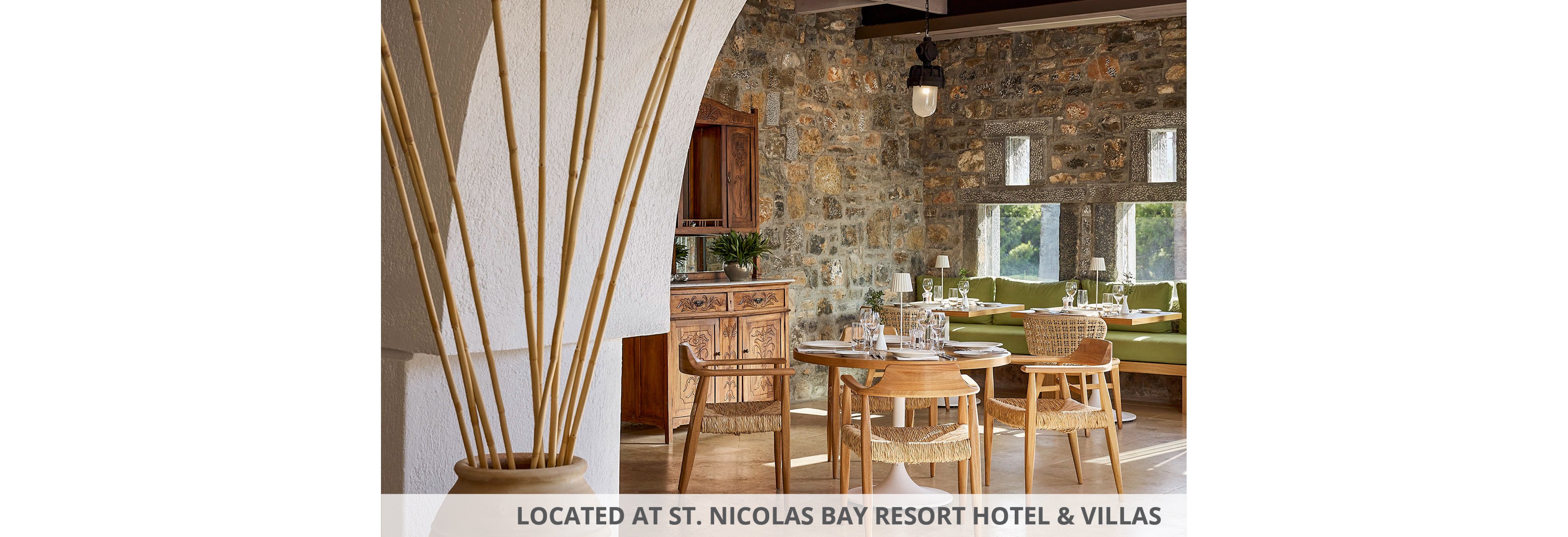 A warm, rustic restaurant interior features wooden tables and woven chairs arranged neatly in a sunlit dining area. Stone walls and a wooden sideboard add texture and character to the space, while a large white arch frames the view. Green cushioned bench seating lines the back wall, complemented by simple table settings and glassware. Natural light streams through rectangular windows, highlighting the earthy tones and relaxed atmosphere.
