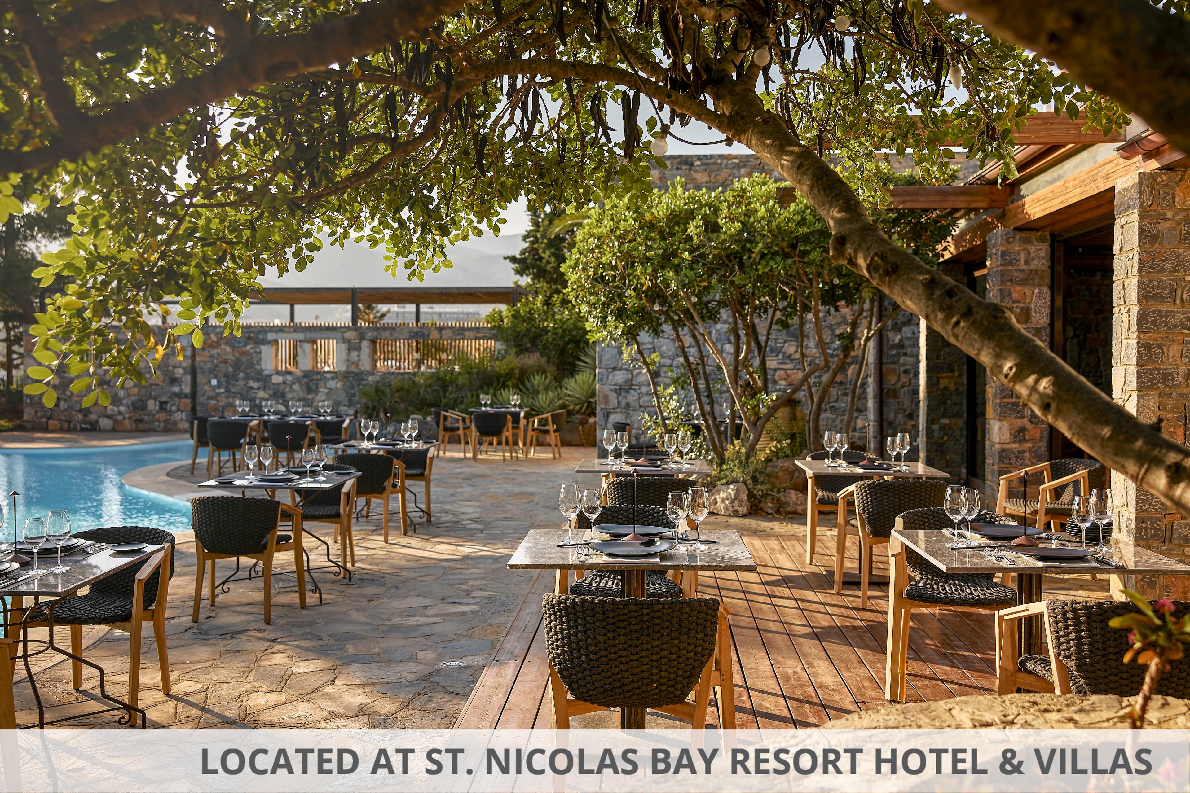 An upscale outdoor restaurant is set beside a curved swimming pool, with neatly arranged tables and chairs on a stone and wooden terrace. Each table is prepared with glassware and place settings, ready for guests. Mature trees and lush greenery provide shade and a natural canopy around the seating area. Warm sunlight highlights the stone walls and wooden structures, creating a relaxed yet sophisticated atmosphere.