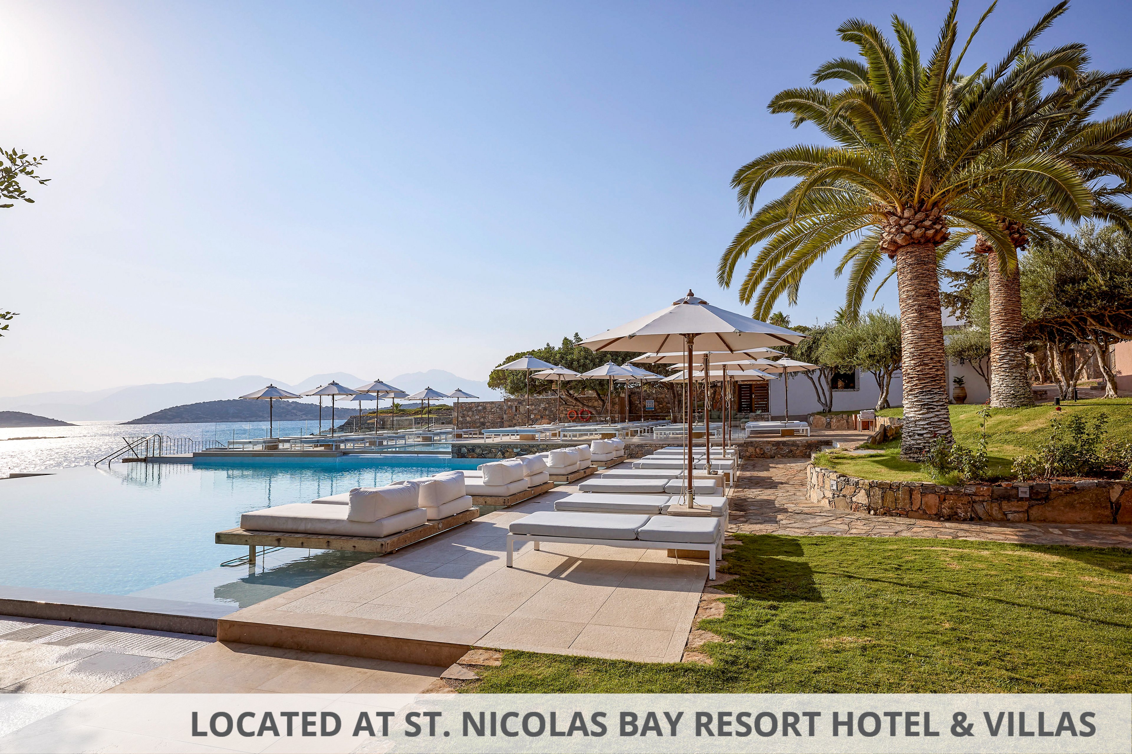 A serene infinity pool overlooks a calm seaside landscape with distant mountains. White sun loungers and umbrellas line the poolside terrace, creating a luxurious resort atmosphere. Palm trees and manicured lawns frame the scene under a clear blue sky. The tranquil setting suggests a relaxing vacation destination by the water.
