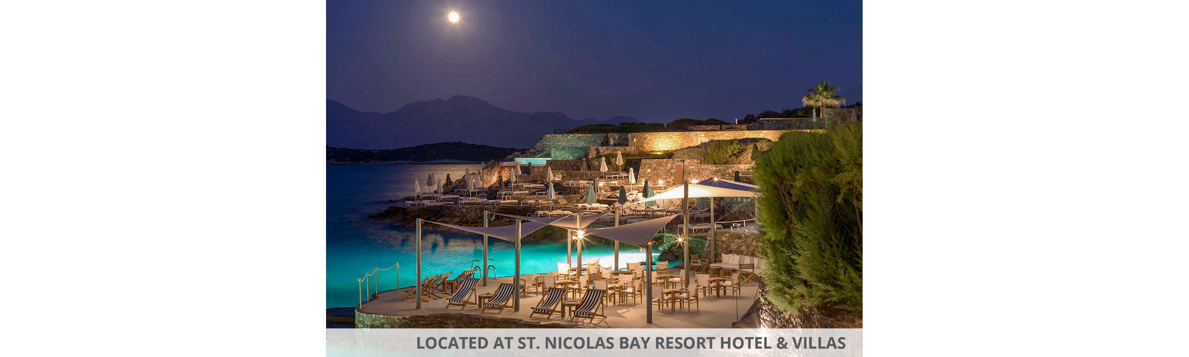 A coastal resort terrace overlooks calm turquoise water under a bright full moon. Lounge chairs and dining tables are arranged neatly on a stone platform by the sea, with sun umbrellas folded for the evening. Warm artificial lights illuminate the seating area and nearby stone walls, contrasting with the cool blue tones of the water and sky. Distant mountains form a dark silhouette on the horizon, enhancing the tranquil nighttime atmosphere.