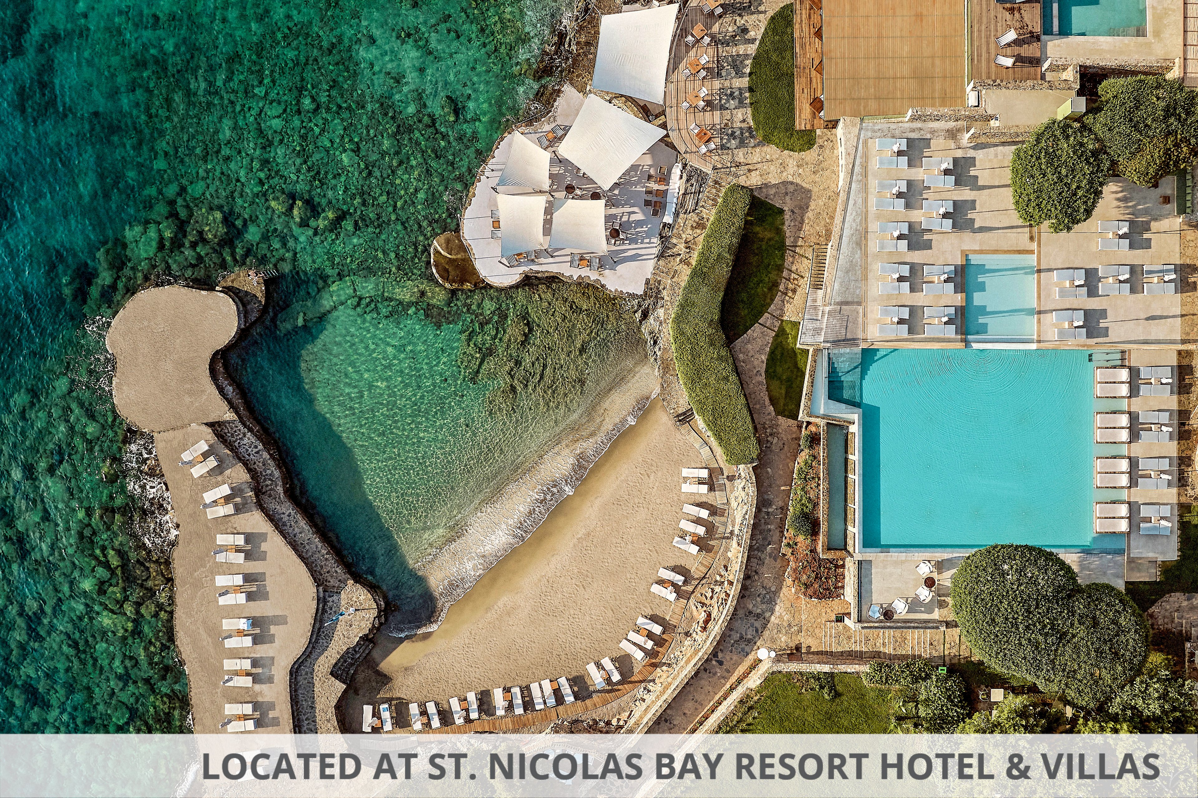 An aerial view captures a luxury seaside resort with a large rectangular swimming pool and adjacent sun loungers. Below the pool area, a curved sandy cove with neatly arranged beach chairs leads into clear turquoise water. White shade structures and stone pathways connect the different terraces and relaxation zones. The scene highlights the contrast between the blue sea, the pool, and the surrounding greenery, emphasizing a tranquil vacation atmosphere.