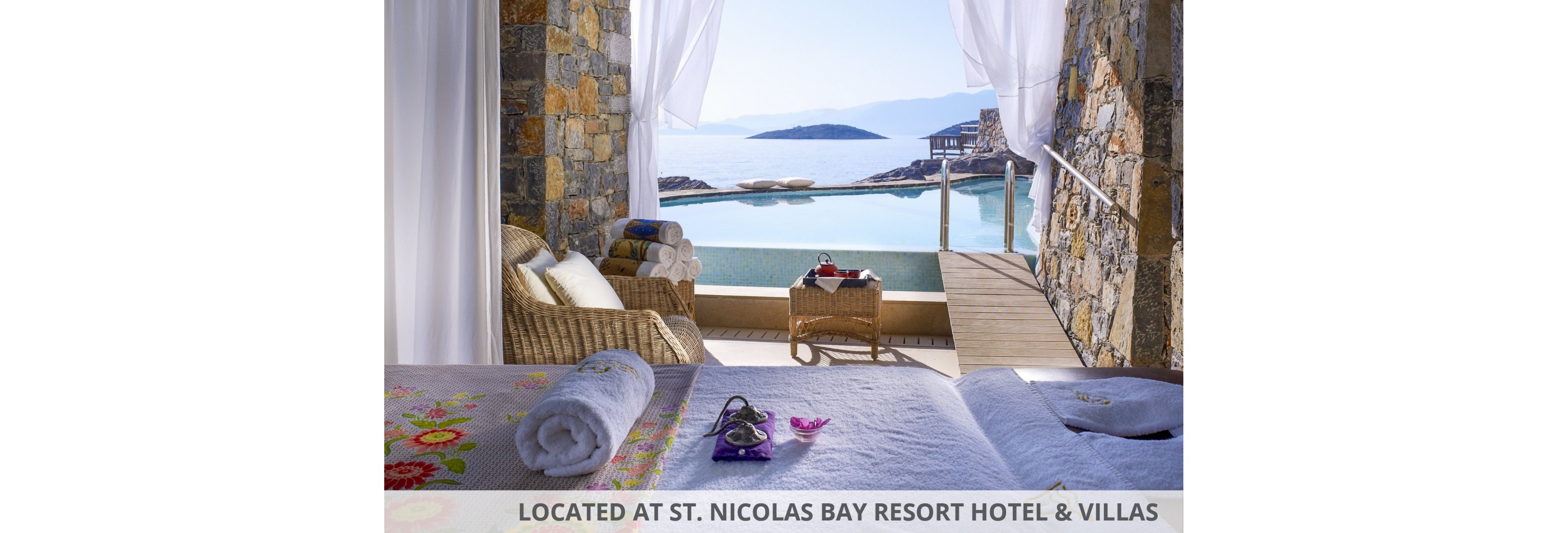 A serene spa suite opens onto a private terrace overlooking a calm sea and infinity pool. A massage bed with floral linens and neatly rolled towels sits in the foreground, suggesting relaxation and wellness treatments. Wicker chairs and a small table with refreshments enhance the cozy, resort-like atmosphere. Soft curtains frame the stone walls and bright natural light, creating a tranquil coastal retreat.