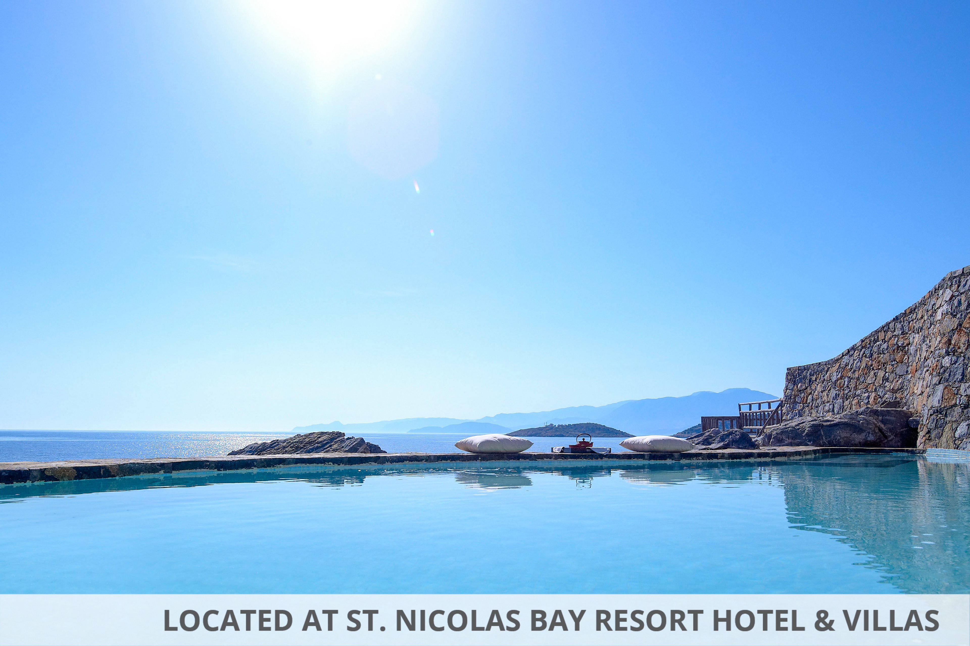 A tranquil infinity pool stretches toward a calm blue sea under a clear sky. The sun shines brightly, reflecting on the still water and creating a peaceful atmosphere. A stone wall lines one side of the pool, adding a rustic architectural element to the scene. Distant mountains and coastline are visible on the horizon, enhancing the sense of openness and relaxation.