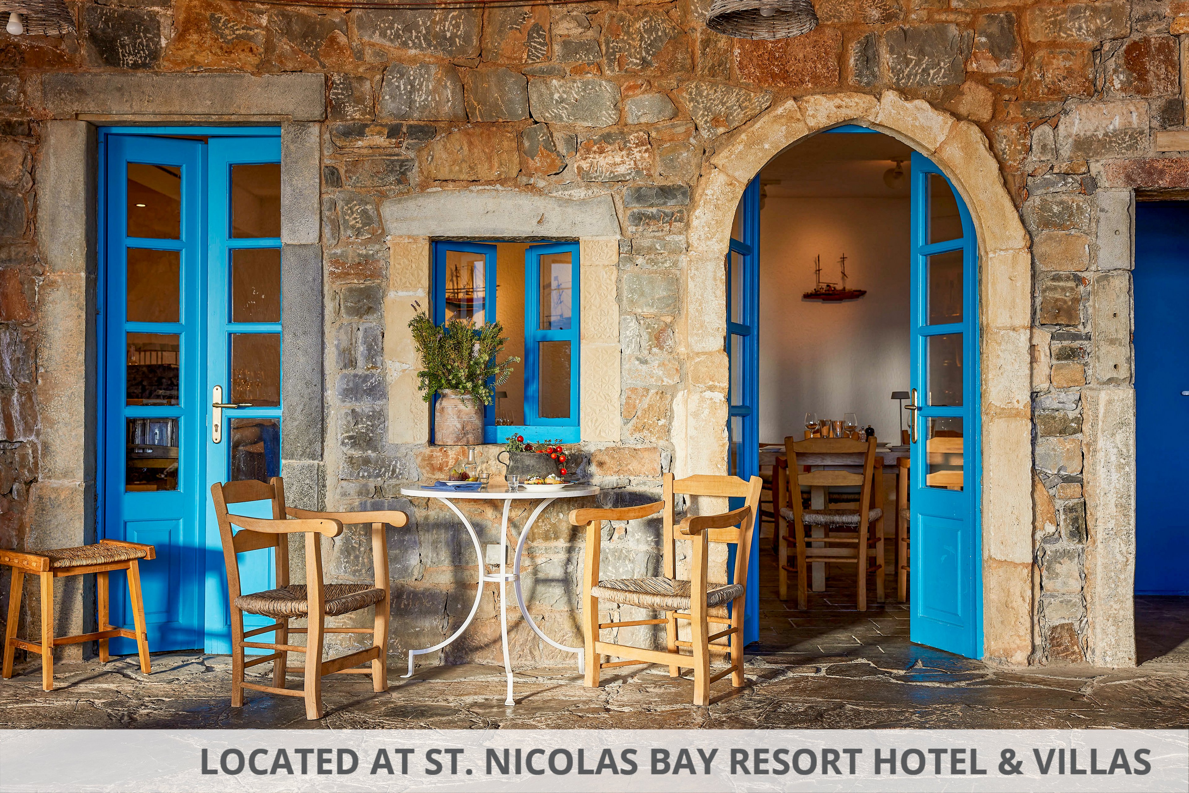 A small outdoor cafe table with wooden chairs sits in front of a rustic stone building. Bright blue doors and window frames contrast with the warm stone walls, creating a Mediterranean feel. Simple wooden furniture and potted flowers add a welcoming touch to the quiet scene. The interior visible through the doorway suggests a relaxed dining area inside.