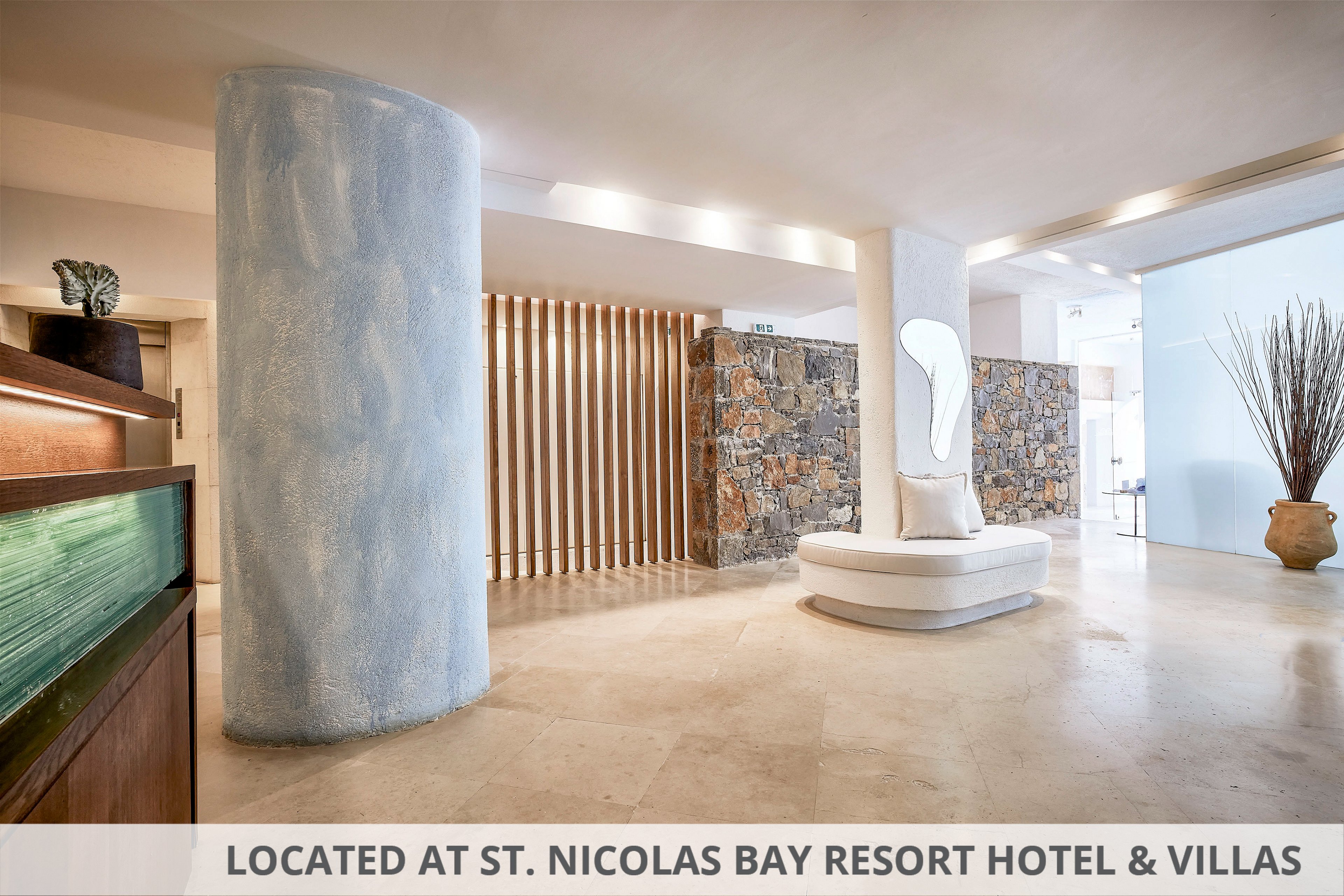 A spacious modern lobby features a large pale blue column and a central white circular seating area. Natural stone walls and vertical wooden slats add texture and warmth to the interior. A sculptural mirror or light element stands above the seating, while a tall vase with dried branches decorates the background. Soft neutral tones and clean lines create a calm, minimalist atmosphere.