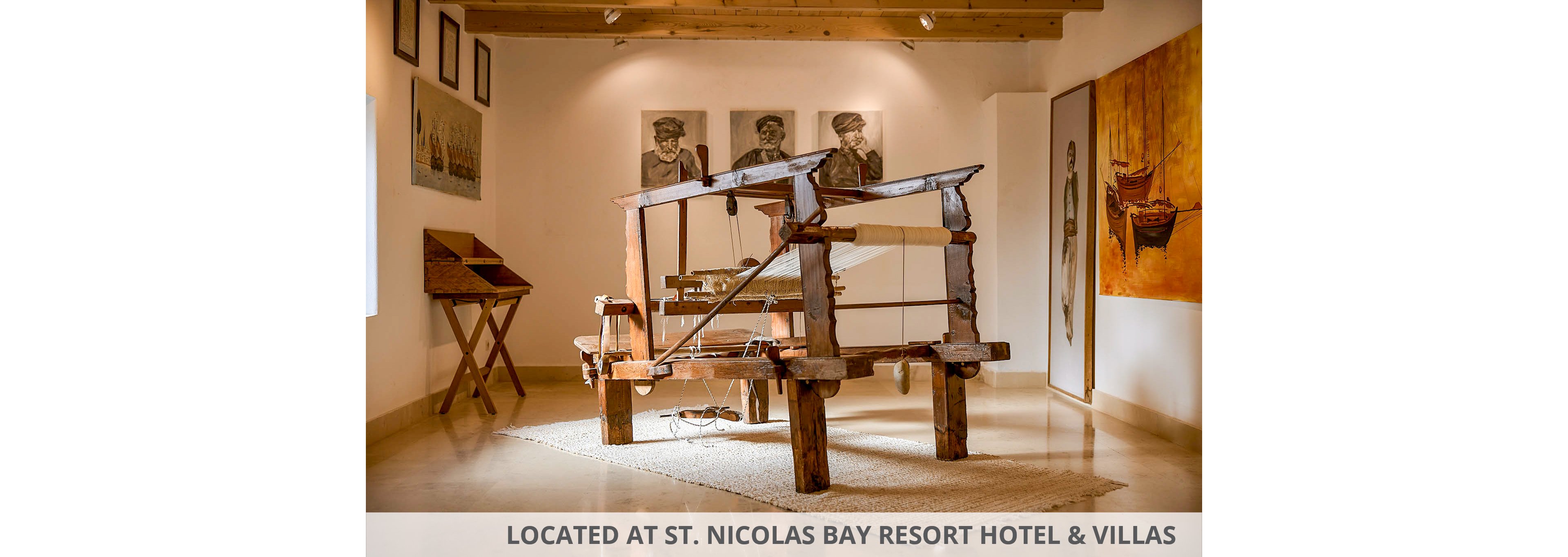 A large traditional wooden loom stands prominently in the center of an art gallery. The room features polished floors, a neutral rug beneath the loom, and wooden beams on the ceiling. Framed portraits and paintings hang on the walls, including images of people and boats. Soft, warm lighting highlights the craftsmanship of the loom and the surrounding artworks, creating a calm, museum-like atmosphere.