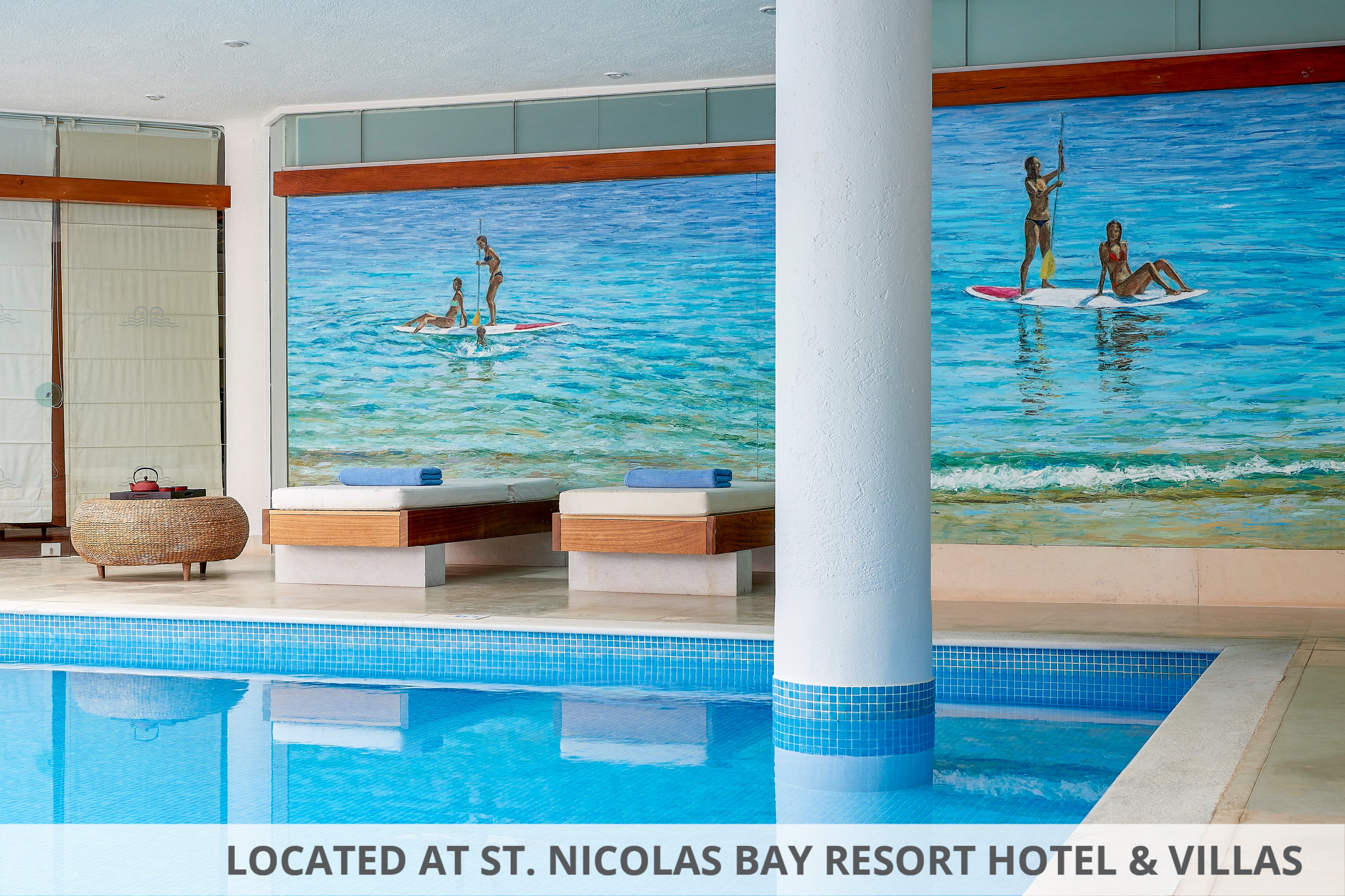 A serene indoor swimming pool features bright blue water and clean modern architecture. Along the far wall, large murals depict people paddleboarding on a calm ocean. Two cushioned daybeds sit beneath the artwork, inviting relaxation beside the pool. A white column and tiled pool edge add geometric structure to the tranquil scene.