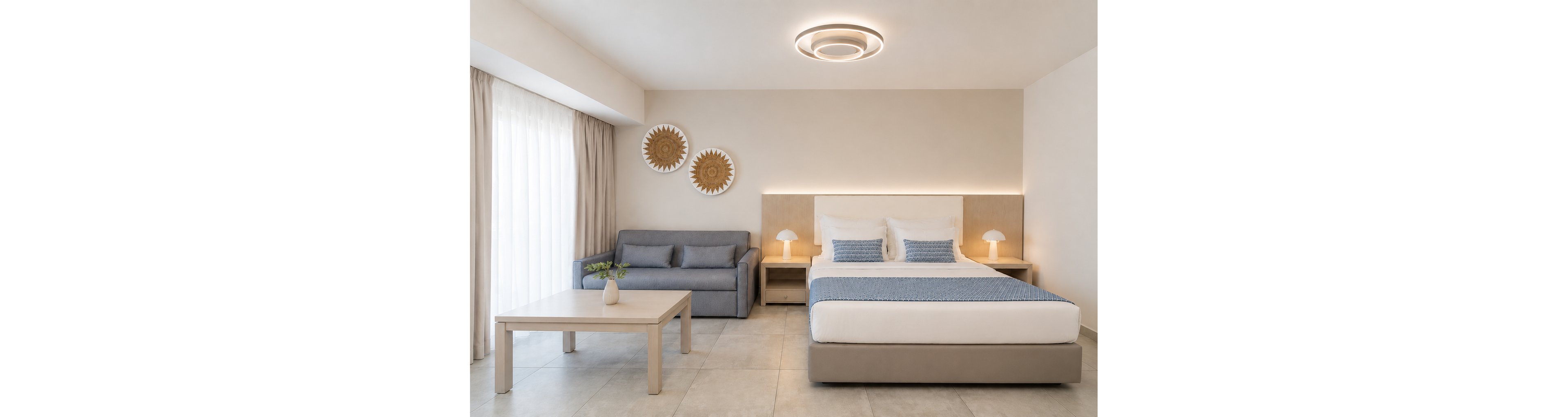 A bright, minimalist bedroom features a large bed with white linens and a light blue textured throw. To the left, a gray sofa and simple wooden coffee table create a small sitting area near floor-to-ceiling curtains. Two round woven wall decorations hang above the sofa, adding natural texture to the neutral space. Soft ambient lighting from a modern ceiling fixture and two bedside lamps enhances the calm, contemporary atmosphere.