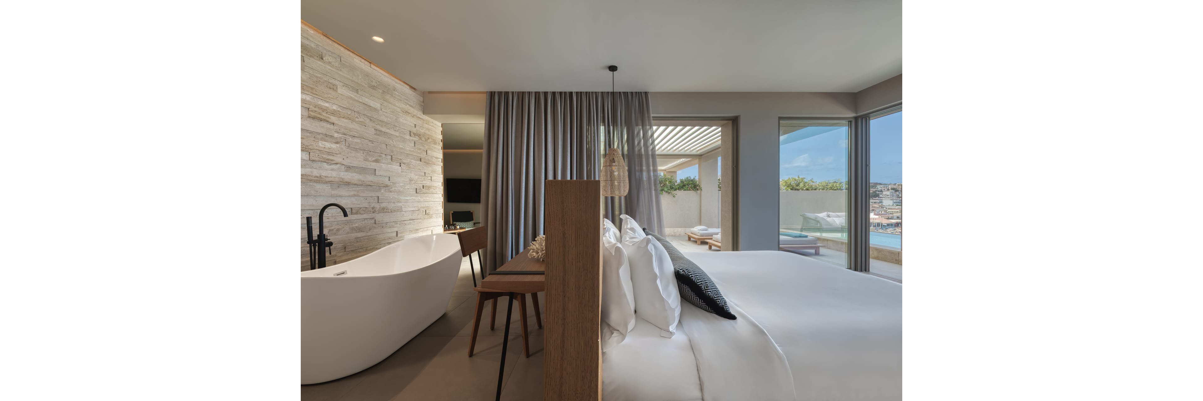 Contemporary hotel suite featuring a white freestanding bathtub and a large bed with crisp white linens. The room includes a wooden desk and chair, stone accent wall, and floor-to-ceiling curtains. Sliding glass doors open to a terrace with outdoor seating and city or coastal views. Soft natural light and neutral tones create a calm, luxurious atmosphere.