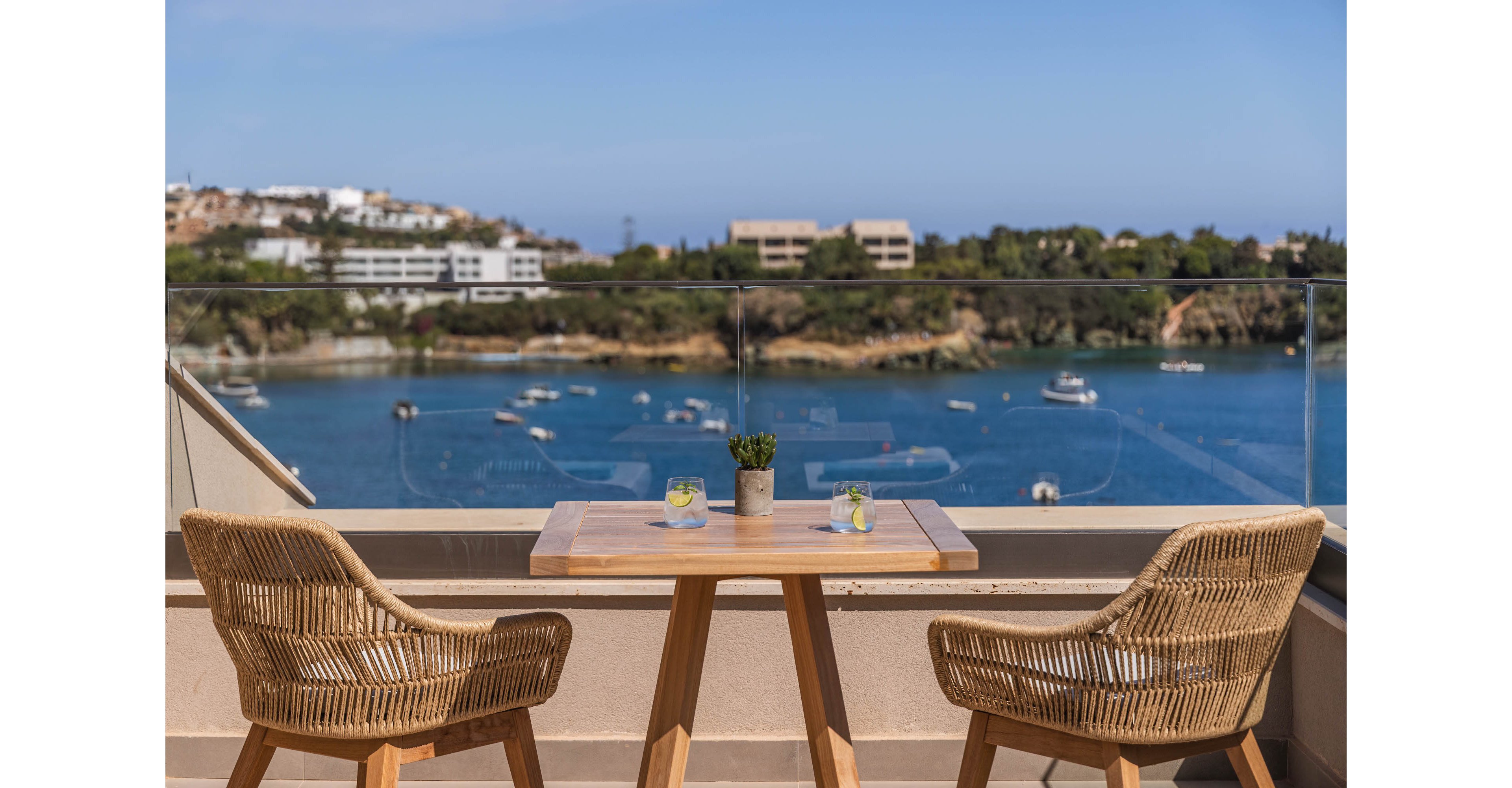A small wooden table with two wicker chairs sits on a terrace overlooking a calm blue bay. Two glasses with drinks and a small potted plant are placed neatly on the table. Beyond the glass railing, boats float on the water and low-rise buildings line the distant shore. The scene suggests a peaceful outdoor seating area with a scenic coastal view under clear skies.