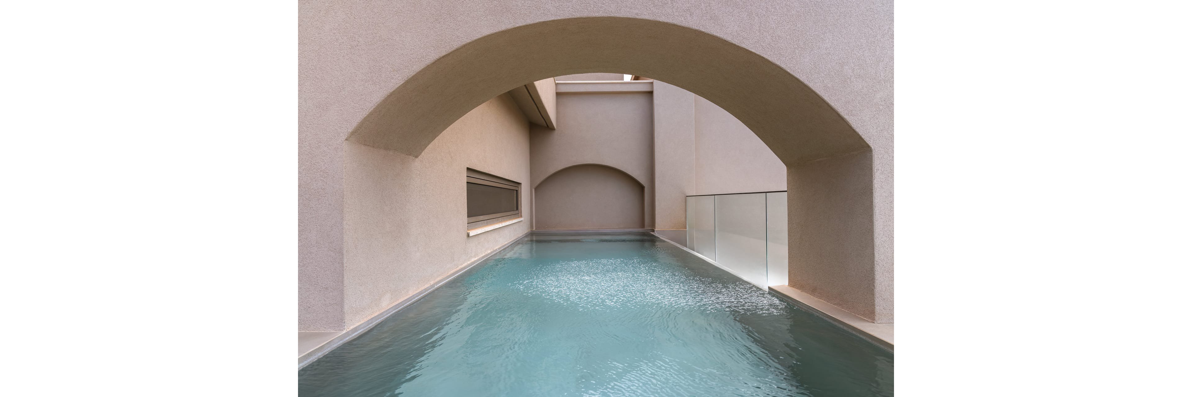 A narrow, modern pool stretches beneath smooth concrete arches in a minimalist architectural space. The water surface is calm with gentle ripples, reflecting the soft neutral tones of the surrounding walls. Glass railing on one side adds a contemporary touch and emphasizes clean lines and symmetry. The overall scene feels quiet, refined, and serene, highlighting the interplay of geometry, light, and water.