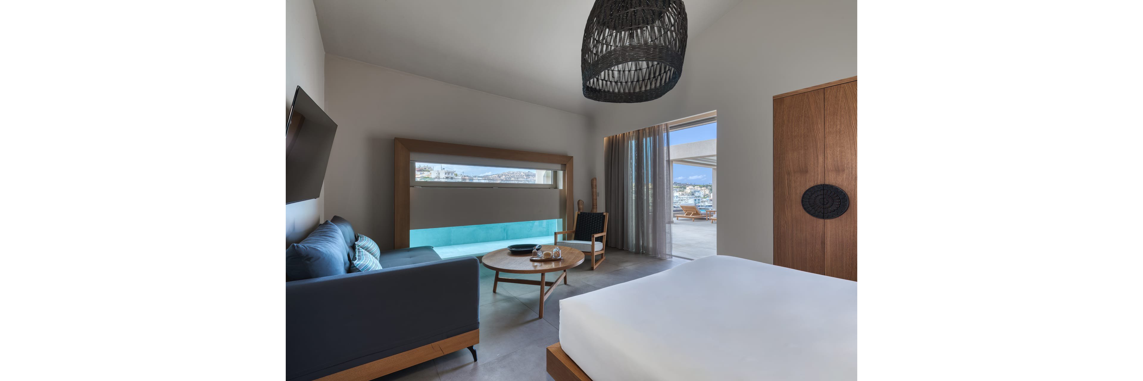 Contemporary hotel room features a large bed, minimalist wooden furniture, and a built-in window aquarium or pool view. A sofa and armchairs surround a round coffee table, creating a relaxed seating area. Floor-to-ceiling curtains open to a terrace with outdoor seating and a scenic coastal city view. Neutral tones, clean lines, and natural light give the space a calm, modern atmosphere.