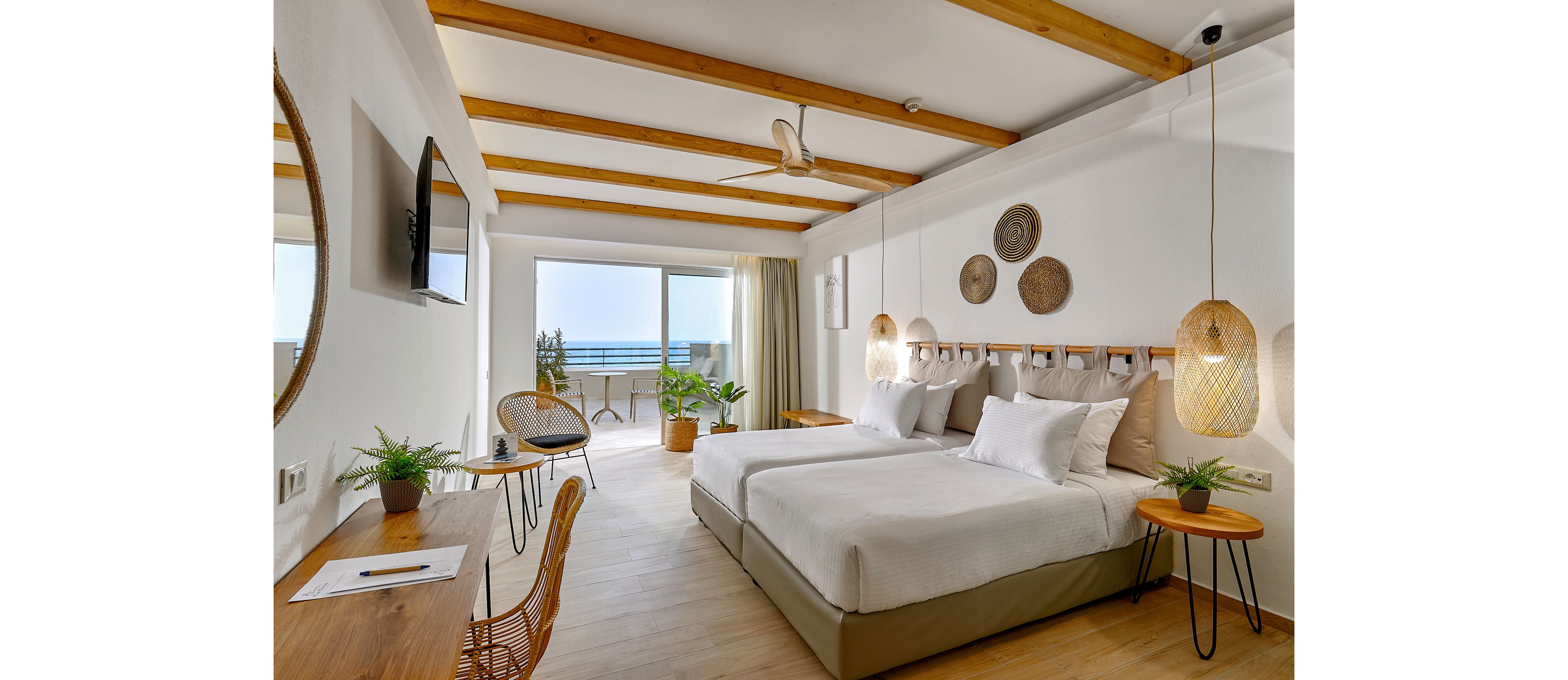 A modern hotel room features two neatly made twin beds with white linens and woven pendant lights. Natural wood accents, wicker chairs, and decorative wall baskets create a warm, coastal style. A desk with plants and a mounted television line one wall, while large glass doors open to a balcony overlooking the sea. Soft daylight fills the space, emphasizing the airy and relaxing atmosphere.