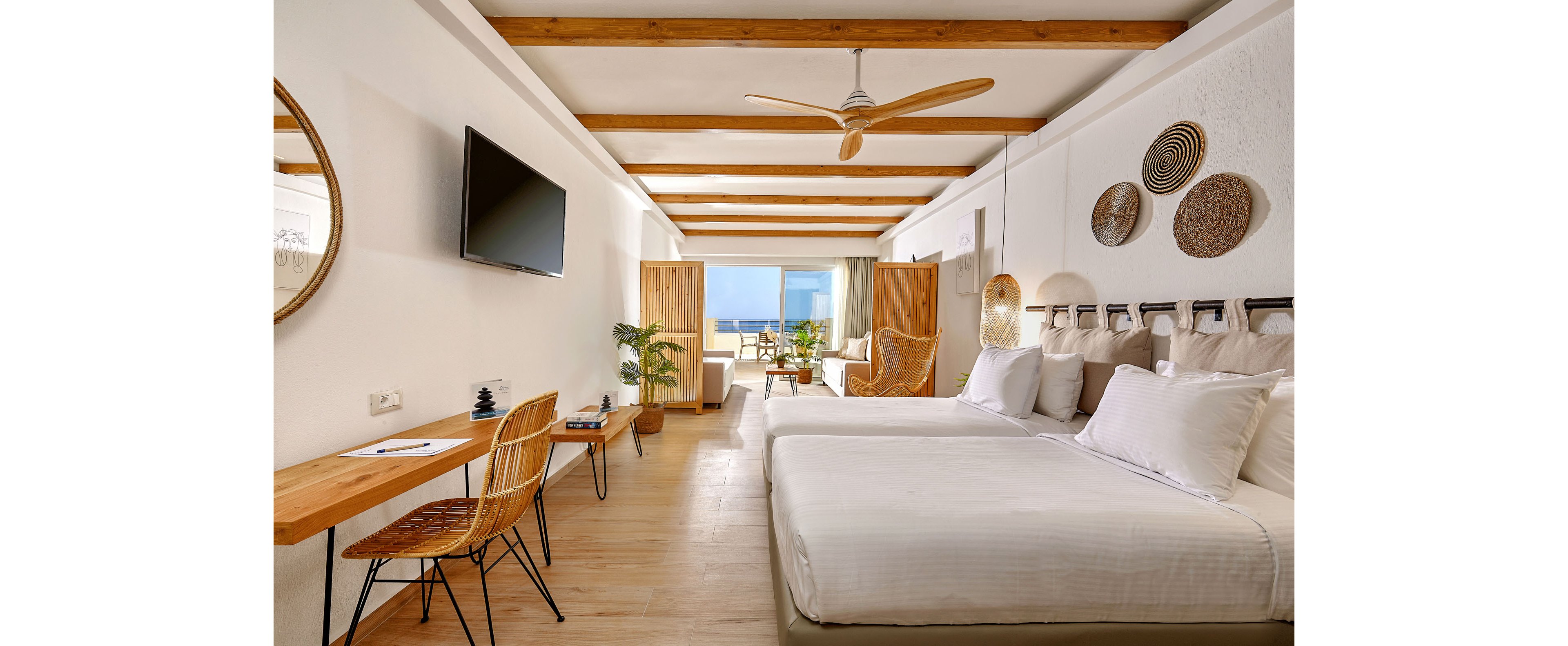 Spacious modern hotel room features two neatly made twin beds with white linens. A wooden desk with a chair, wall-mounted television, and decorative woven wall art enhance the contemporary coastal style. Natural wood beams, ceiling fan, and indoor plants add warmth to the bright white interior. Large glass doors open to a balcony with seating and a clear view of the ocean in the distance.