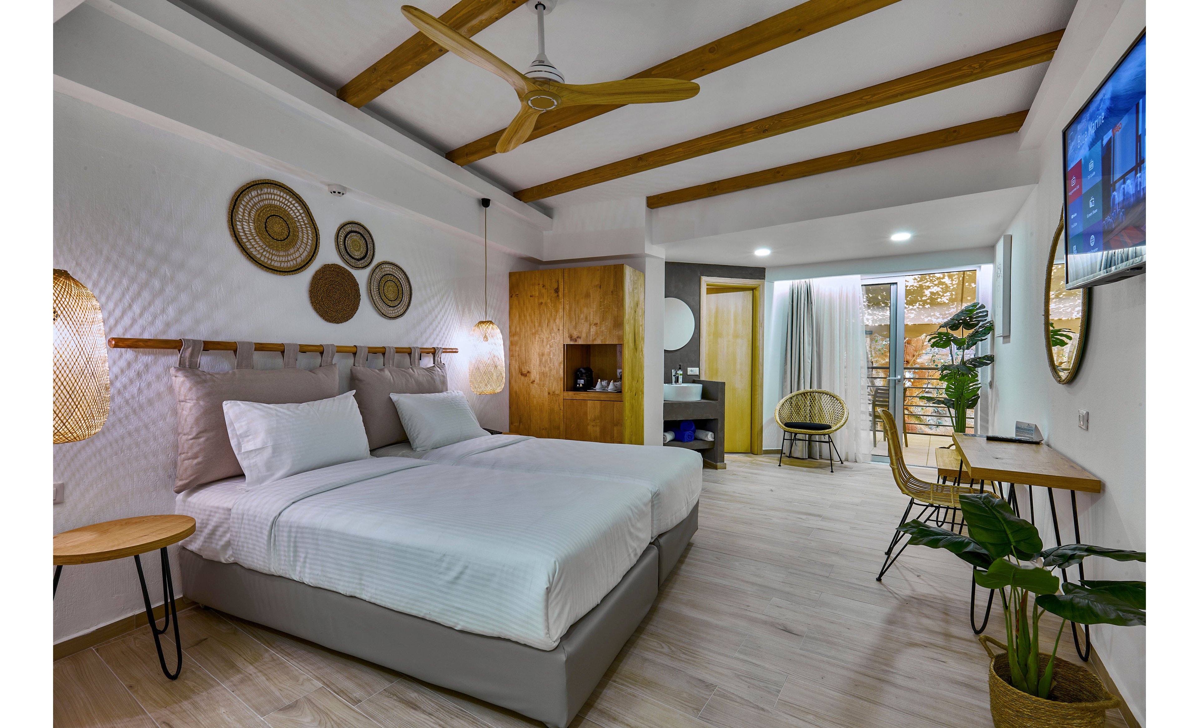 Contemporary hotel room features a large double bed with white linens and decorative wall art above the headboard. The space includes wooden ceiling beams, a ceiling fan, and warm pendant lighting that create a cozy atmosphere. A small seating area with wicker chairs and a desk faces a wall-mounted television, while potted plants add a touch of greenery. Floor-to-ceiling curtains open to a balcony with outdoor seating and a view of trees, enhancing the airy, vacation feel.